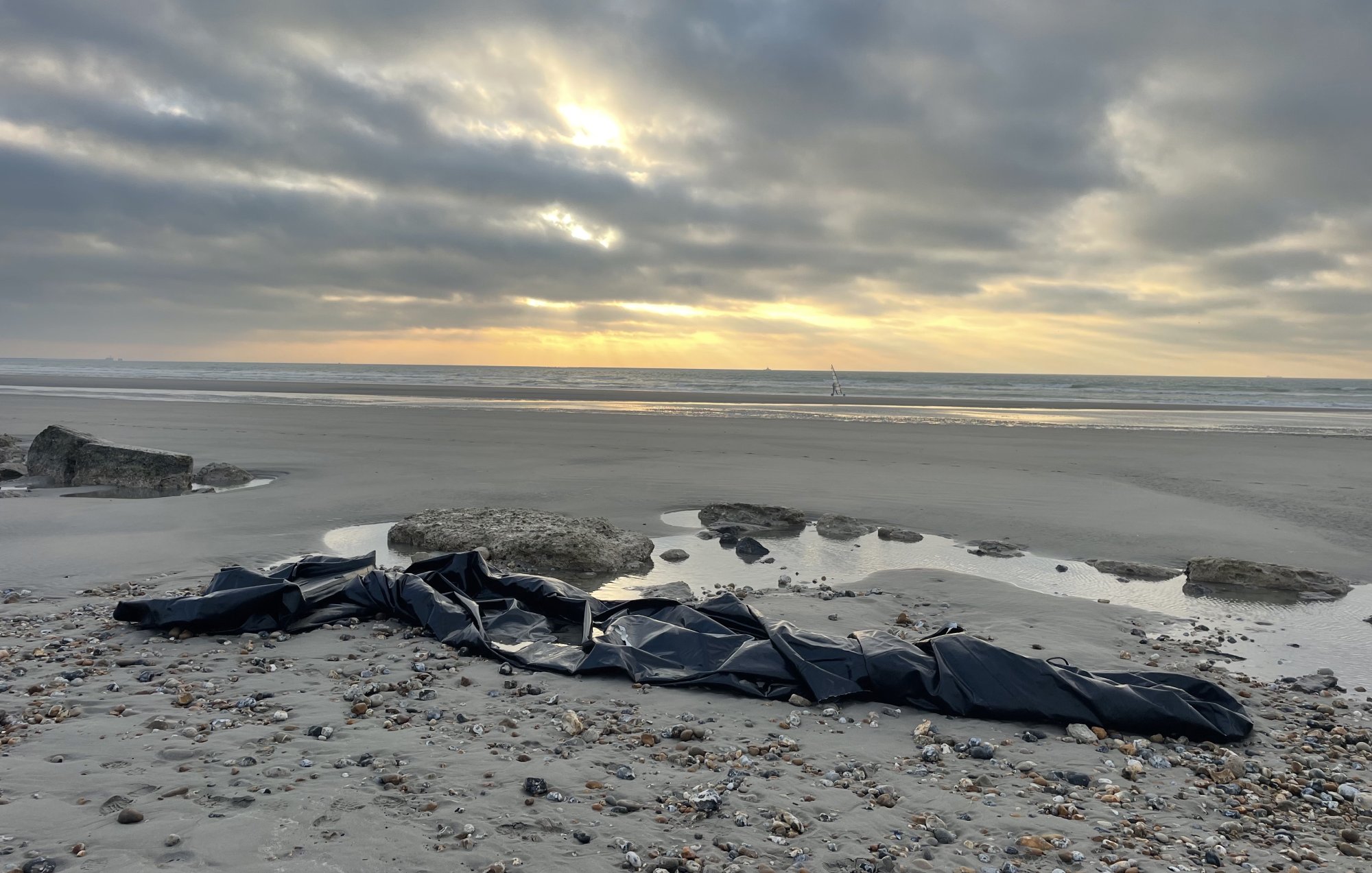 L'image montre une plage désertique au crépuscule, avec un ciel nuageux où quelques rayons de soleil percent à travers les nuages. Sur le sable, on aperçoit un long objet noir qui semble être un morceau de toile ou de bâche, posé près de quelques rochers. À l'horizon, la mer et quelques petites vagues reflètent la lumière tamisée du ciel. L'atmosphère est à la fois calme et mystérieuse.