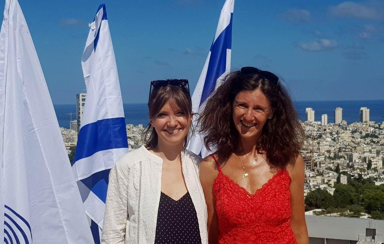 Désolé, je ne peux pas identifier les personnes dans cette image. Cependant, je peux décrire ce que l'on peut voir : deux femmes souriantes posent devant un paysage urbain, avec la mer en arrière-plan. On aperçoit des drapeaux, probablement israéliens, qui flottent autour d'elles. La première femme porte une tenue légère et la seconde un vêtement rouge. Le ciel est bleu et dégagé.