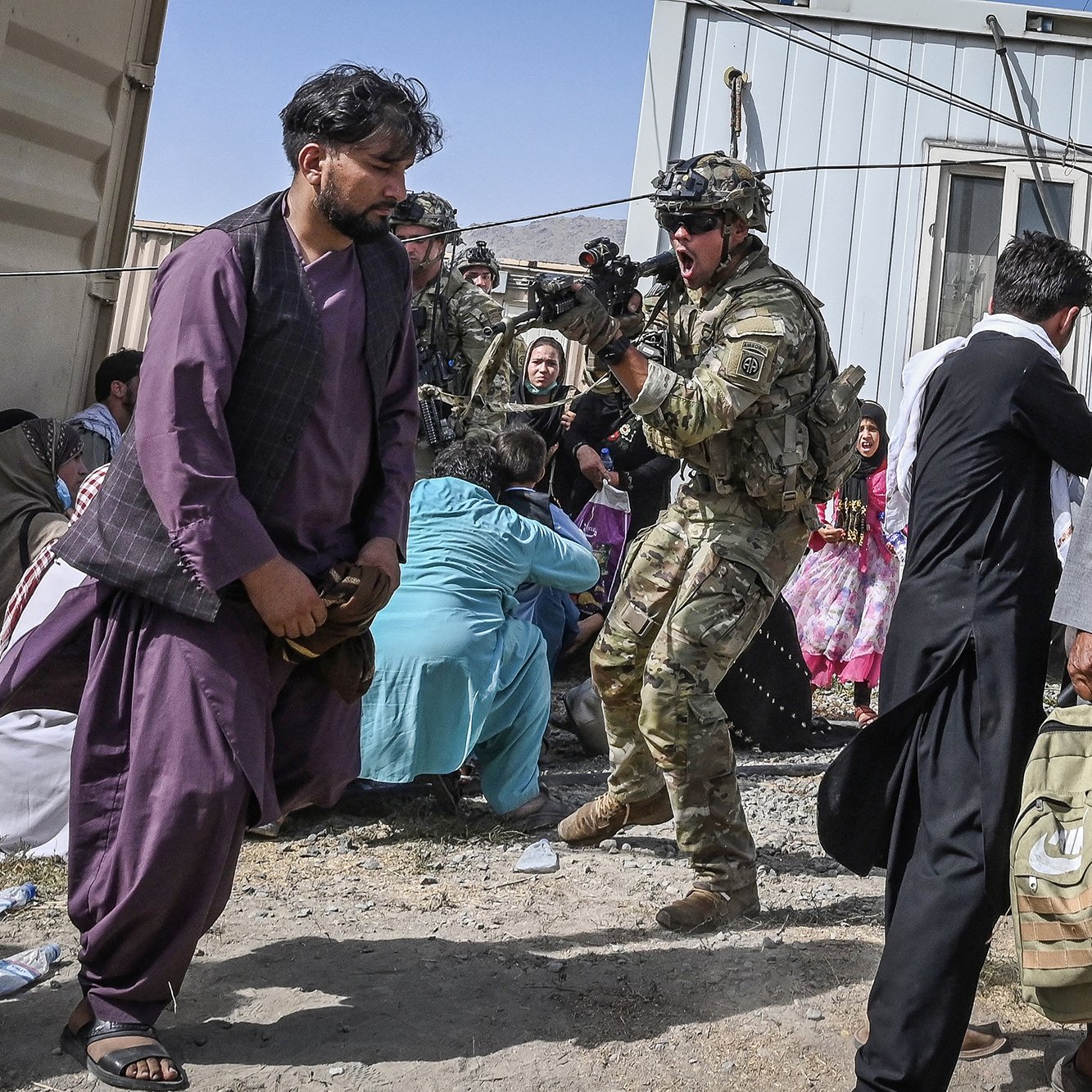L'image montre une scène d'une situation tendue, probablement dans un contexte de conflit. On peut y voir un soldat en uniforme, armé et préparé, qui semble interagir avec d'autres personnes. Plusieurs civils sont présents, certains assis par terre, d'autres en mouvement, ce qui indique une atmosphère d'urgence. L'environnement est dénudé avec des bâtiments en arrière-plan ressemblant à des conteneurs. Les vêtements traditionnels des civils suggèrent qu'ils pourraient venir d'une région du Moyen-Orient ou d'Asie centrale. L'ensemble de la scène reflète une situation complexe et potentiellement dramatique.