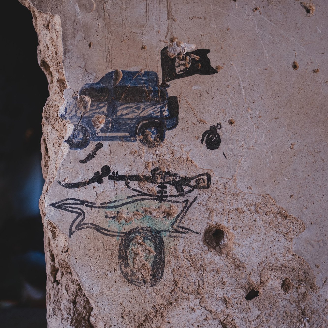 L'image montre un mur en béton décrépit où des graffitis sont visibles. On peut y voir une représentation d'un véhicule, probablement une voiture de police, accompagnée d'un drapeau. En dessous, il y a un dessin stylisé d'un squelette, surmonté d'une forme qui pourrait évoquer un traîneau ou un chariot. Le mur lui-même présente des traces d'usure et des fissures, ajoutant une dimension d'abandon à la scène.