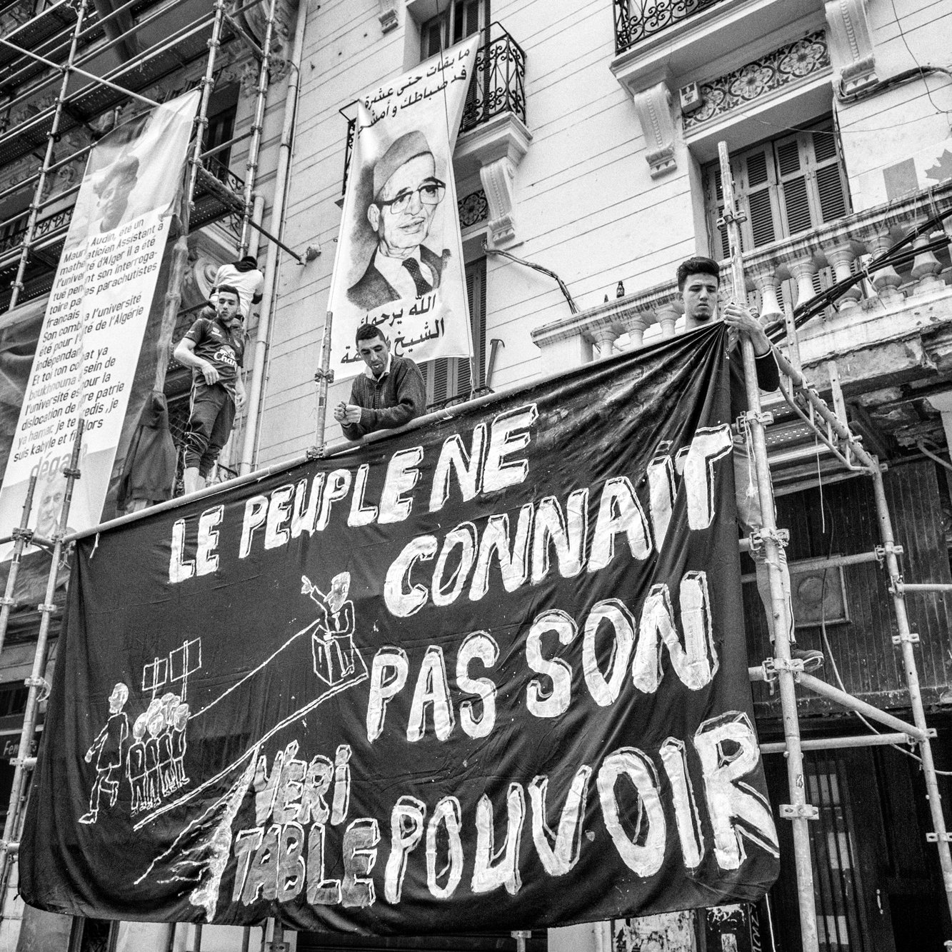 L'image présente une scène en noir et blanc où des personnes sont perchées sur un échafaudage face à un bâtiment. Ils tiennent une grande bannière sur laquelle est écrit en lettres blanches : "LE PEUPLE NE CONNAÎT PAS SON POUVOIR". En arrière-plan, on peut voir des affiches et des motifs typiques d'un environnement urbain. Cette composition évoque un sentiment de protestation ou de revendication sociale.