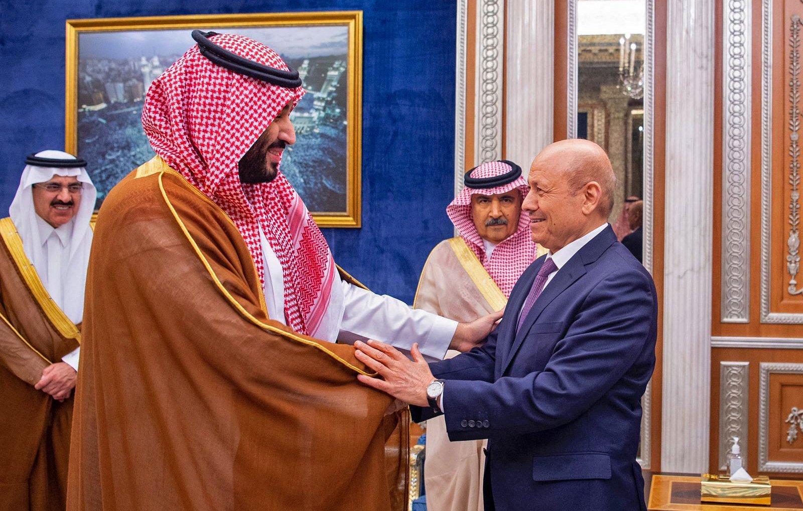 L'image montre deux hommes en costume, engagés dans une interaction amicale, probablement lors d'une rencontre officielle. L'un des hommes porte un habit traditionnel avec un châle. L'arrière-plan présente un décor élégant, typique d'un palais, avec des éléments architecturaux raffinés. D'autres personnes en tenue formelle sont visibles, suggérant un contexte diplomatique ou cérémoniel.