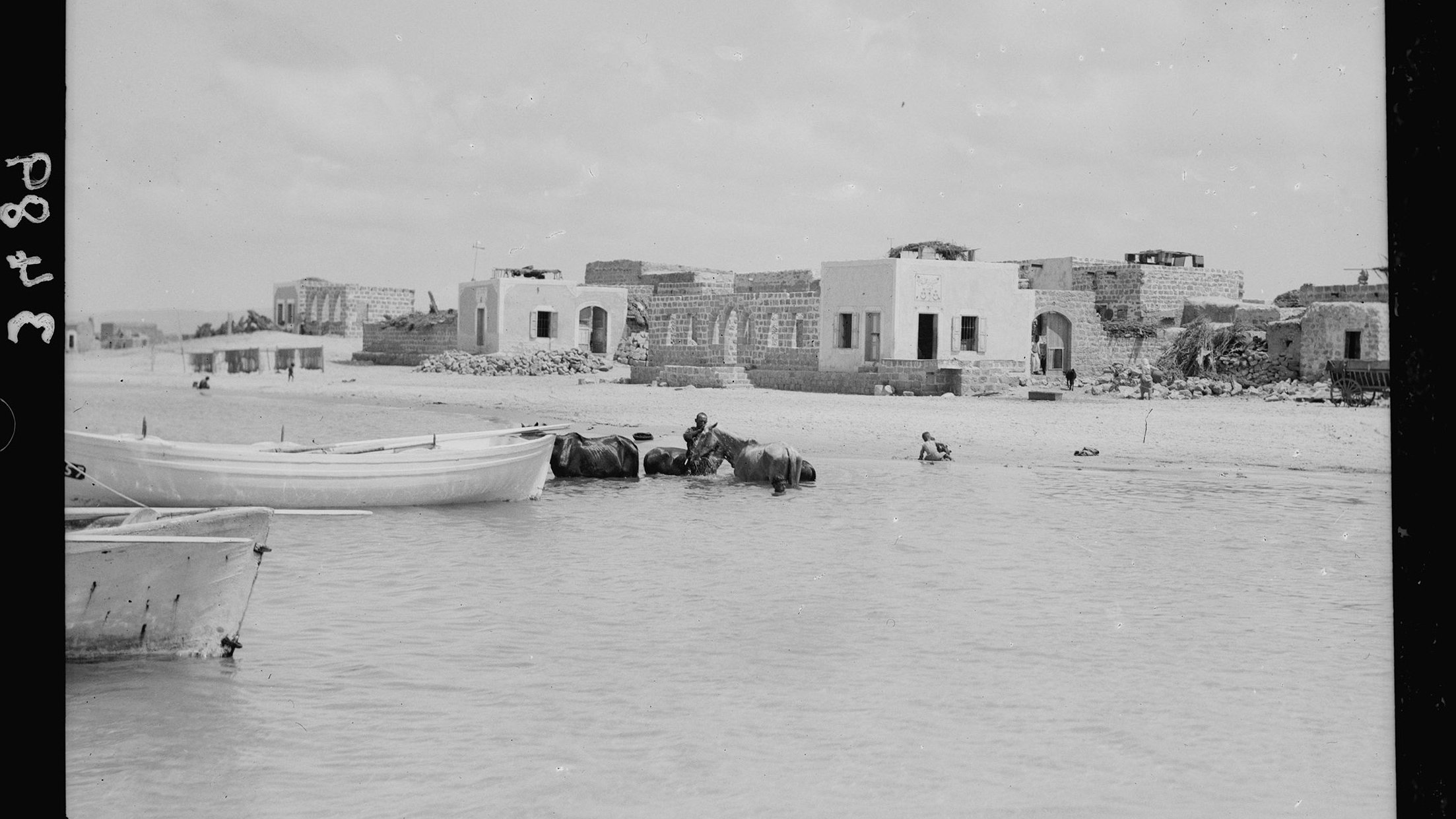 L'image montre une scène maritime où plusieurs bateaux, probablement des barques, sont amarrés au bord d'une eau peu profonde. En arrière-plan, on peut apercevoir des bâtiments en ruines, suggérant une ancienne architecture. À proximité, un homme semble se trouver dans l'eau, tandis qu'un autre est à cheval ou monté sur un animal aquatique. Le ciel est nuageux, apportant une lumière diffuse à l'ensemble de la scène. L'atmosphère semble tranquille et un peu nostalgique, évoquant un passé révolu.