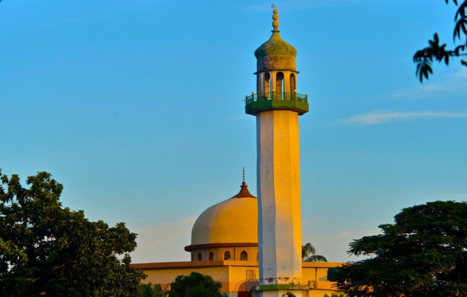 L'image montre un minaret d'une mosquée, qui s'élève majestueusement contre un ciel bleu clair. Le minaret, de couleur claire, présente une colonnade verte au sommet, et un dôme arrondi se trouve à proximité. Des arbres verdoyants entourent la mosquée, ajoutant une touche de nature à la scène paisible. L'ensemble dégage une atmosphère sereine, typique des lieux de culte.