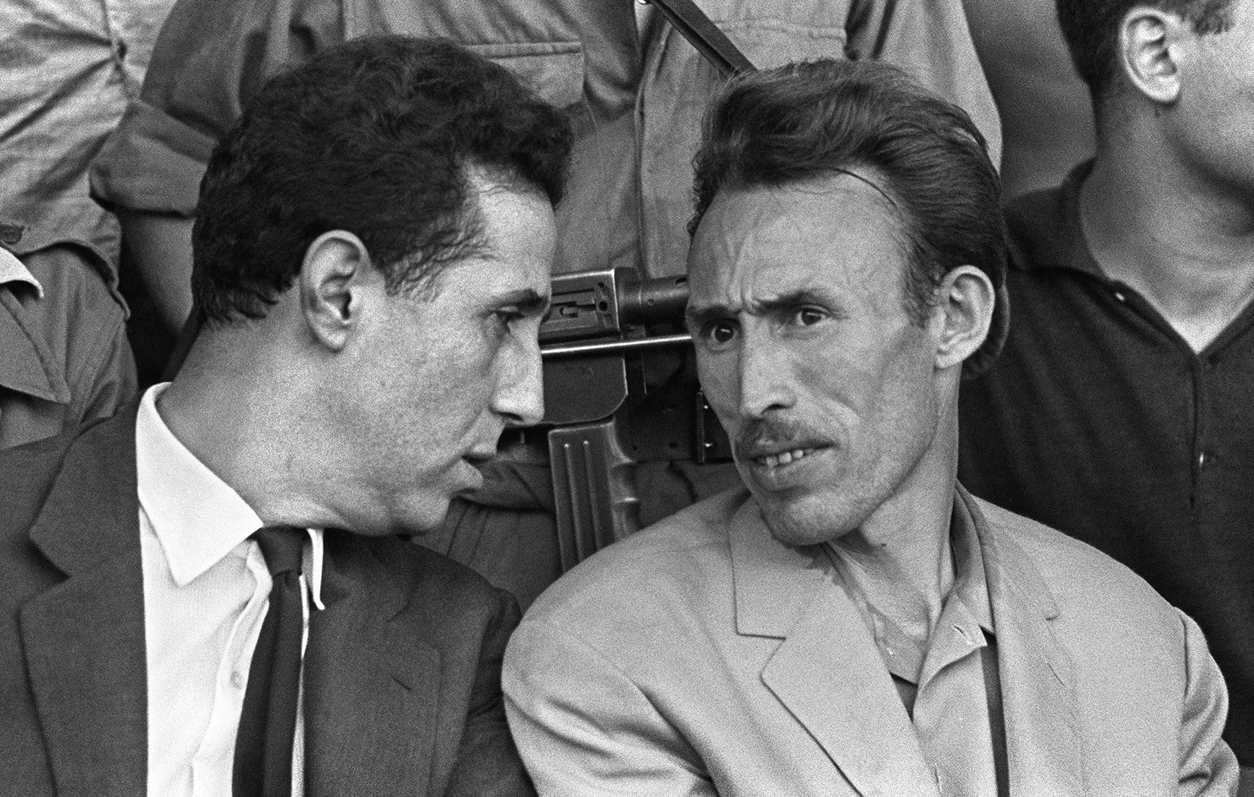 Cette image en noir et blanc montre deux hommes en discussion. L'un porte un costume sombre avec une cravate, tandis que l'autre est habillé de manière plus décontractée avec une veste claire. Ils semblent en train d'échanger des informations ou des réflexions, affichant des expressions attentives. En arrière-plan, on peut apercevoir d'autres personnes, et un fusil est visible, suggérant un contexte de tension ou de sécurité. L'image capte une moment d'intimité entre les deux hommes.