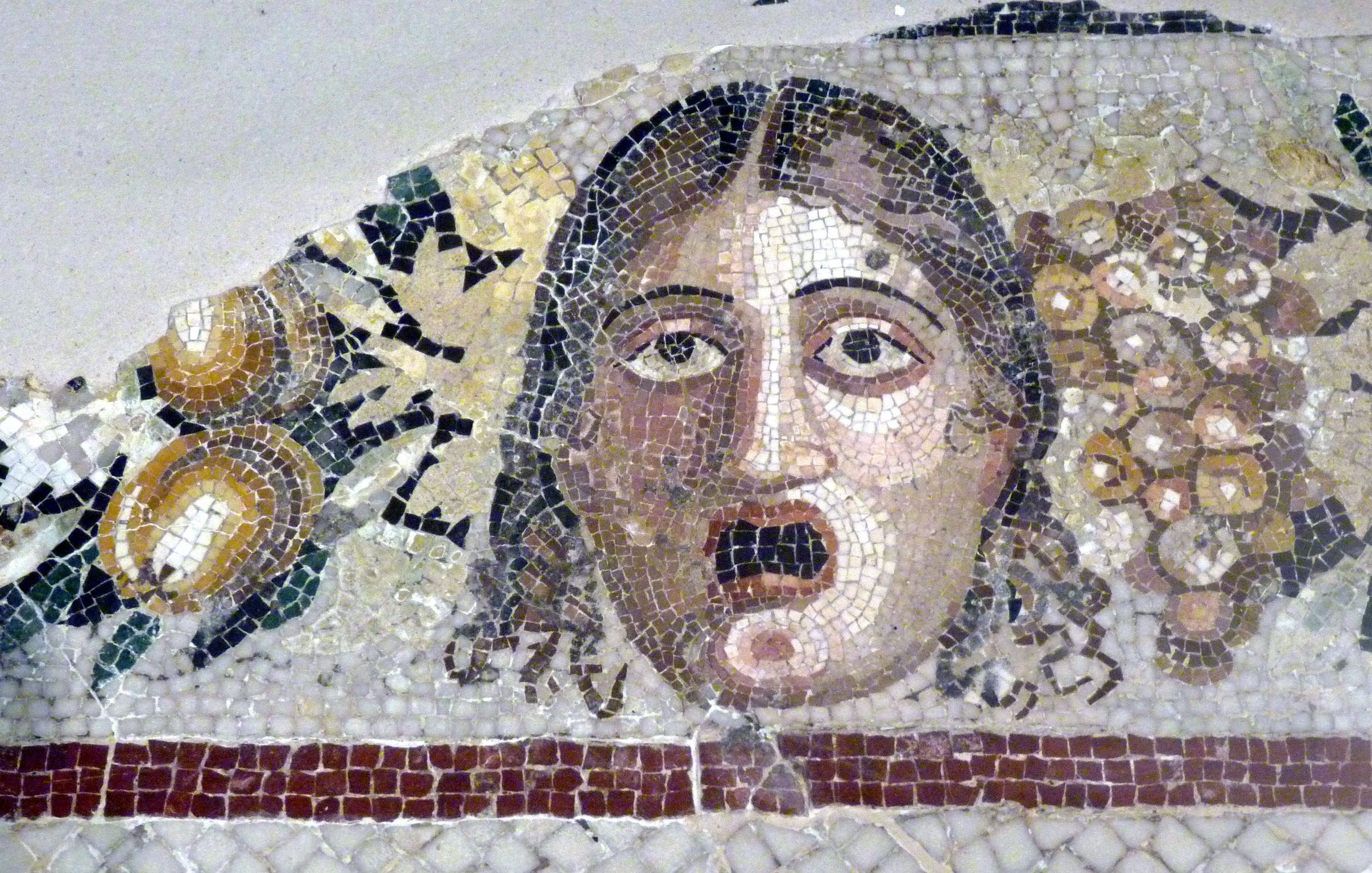 The image depicts a detailed mosaic featuring a face with a startled or shocked expression. The individual's wide eyes and open mouth convey a sense of surprise or fear. Surrounding the face are decorative elements, including clusters of grapes and possibly other fruits or foliage, which suggest a theme of abundance or festivity. The mosaic is composed of various colored tiles, showcasing intricate craftsmanship typical of ancient art.