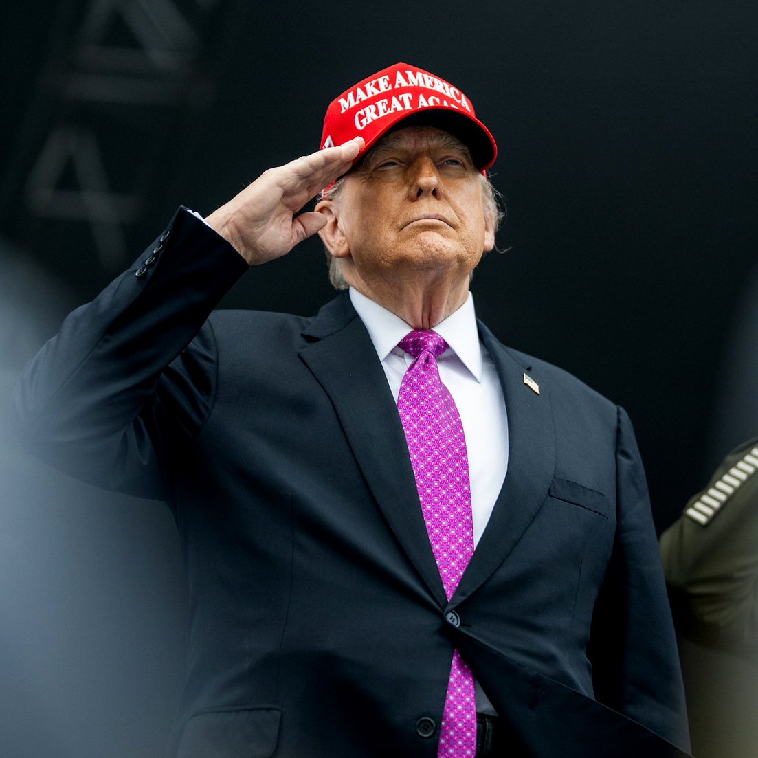 مردی با کلاهی قرمز و پیراهن مشکی، با دست بر پیشانی ایستاده و سلام می‌کند.