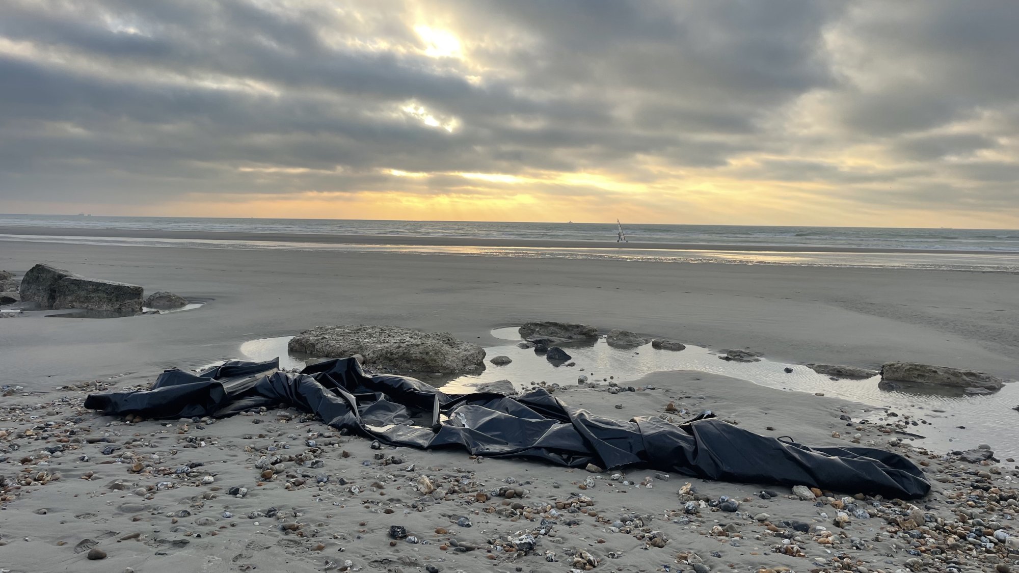 L'image montre une plage désertique au crépuscule, avec un ciel nuageux où quelques rayons de soleil percent à travers les nuages. Sur le sable, on aperçoit un long objet noir qui semble être un morceau de toile ou de bâche, posé près de quelques rochers. À l'horizon, la mer et quelques petites vagues reflètent la lumière tamisée du ciel. L'atmosphère est à la fois calme et mystérieuse.