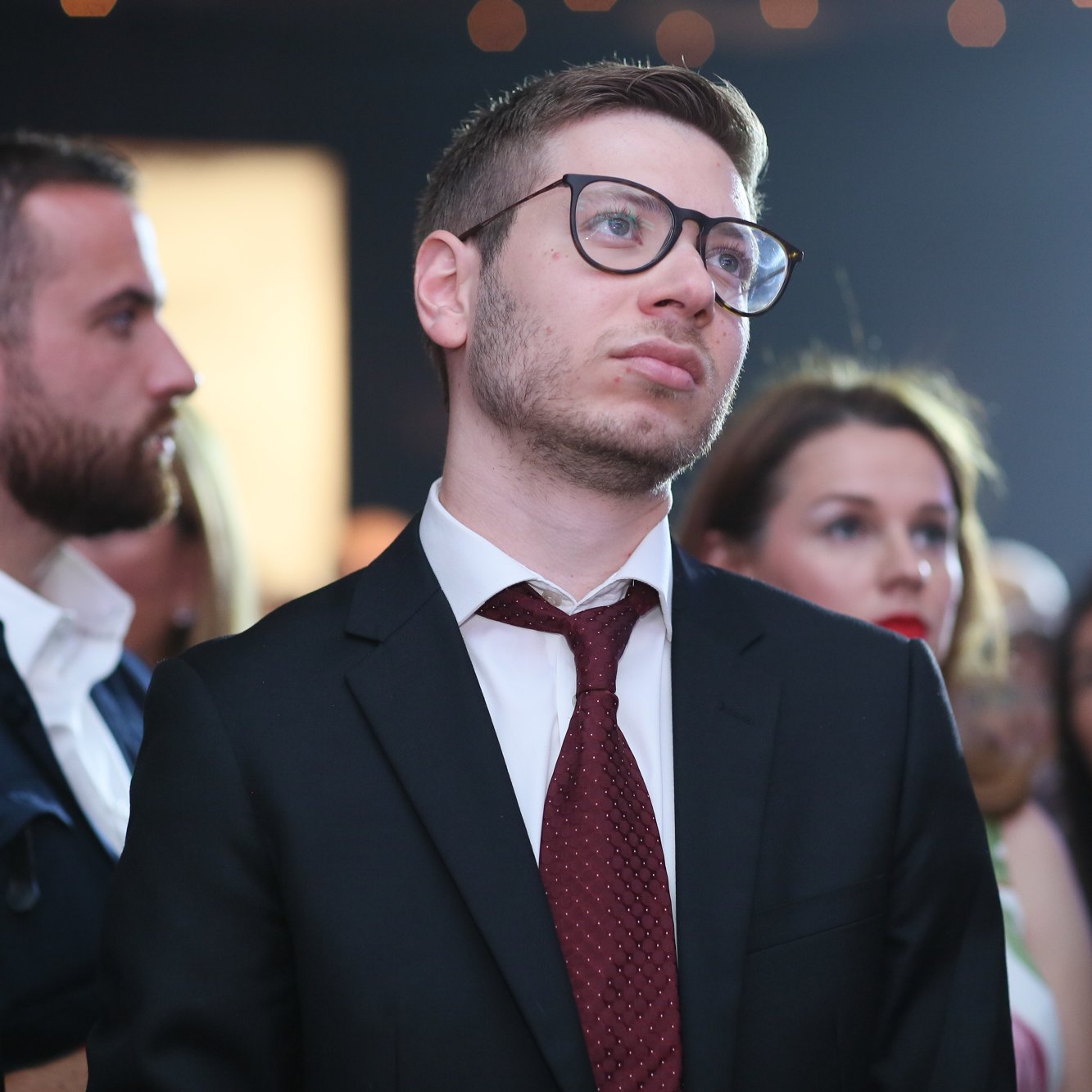 الصورة تُظهر مجموعة من الأشخاص في حدث اجتماعي أو عرض. في المقدمة، يبدو شاب يرتدي بذلة سوداء وقميصًا أبيض ورابطة عنق حمراء، وهو ينظر للأعلى بتعبير تأملي. خلفه يوجد العديد من الأشخاص، بعضهم يرتدي ملابس رسمية وآخرون بملابس أقل رسمية، مما يدل على تنوع الحضور. الإضاءة في الخلفية تعطي جوًا احتفاليًا.