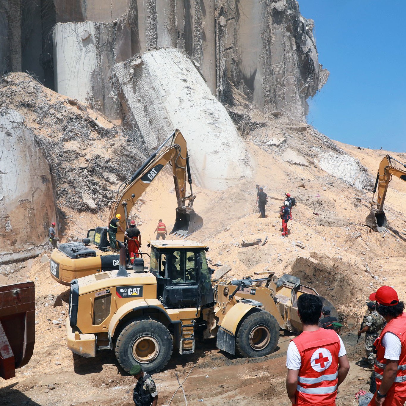 L'image montre un site de construction ou de déblaiement où des engins de chantier, tels que des pelles mécaniques et des bulldozers, sont en action. Au premier plan, des travailleurs en uniforme, y compris des membres de la Croix-Rouge, supervisent les opérations. En arrière-plan, on peut voir des débris et des structures effondrées, suggérant une situation d'urgence ou de nettoyage après un incident. Le ciel est dégagé et ensoleillé, ajoutant un contraste avec l'environnement de travail.