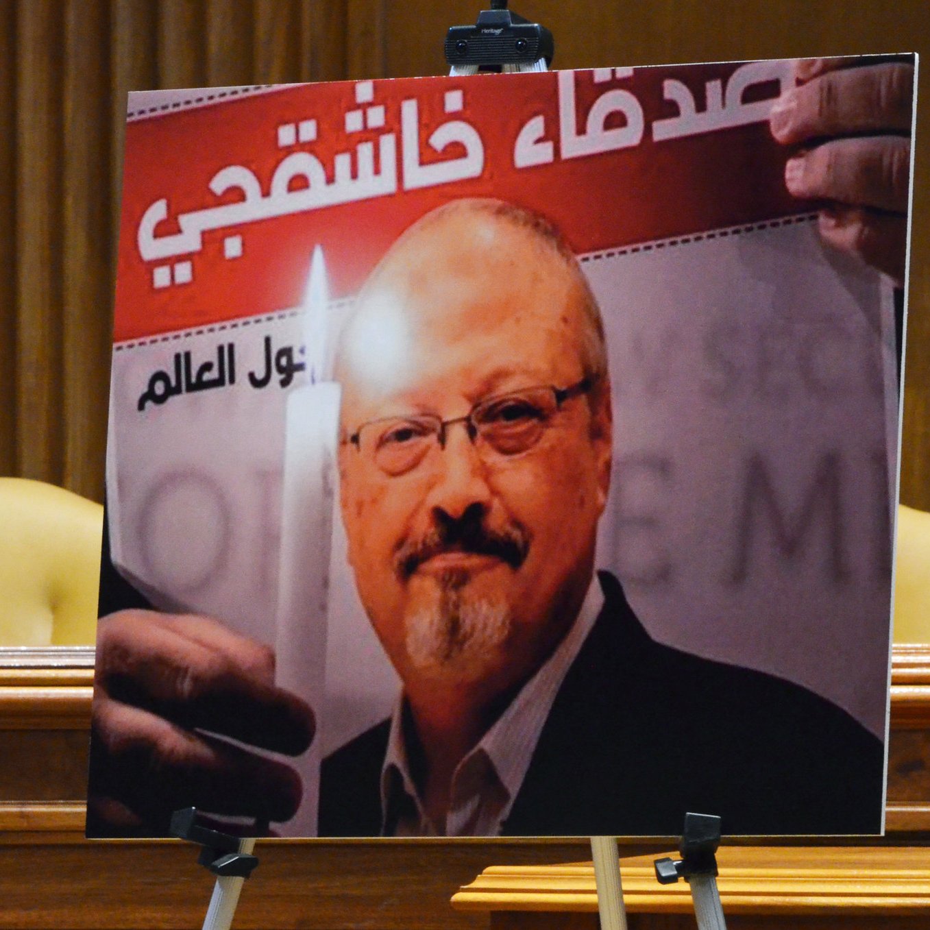 L'image montre un portrait d'un homme tenant une bougie, accompagné d'un texte en arabe et en français. Le fond de l'affiche évoque un hommage ou un soutien, probablement lié à une question de justice. Le contexte global semble être une commémoration ou une manifestation pour défendre les droits humains. Deux chaises sont visibles en arrière-plan, suggérant que l'affiche est présentée lors d'un événement formel.