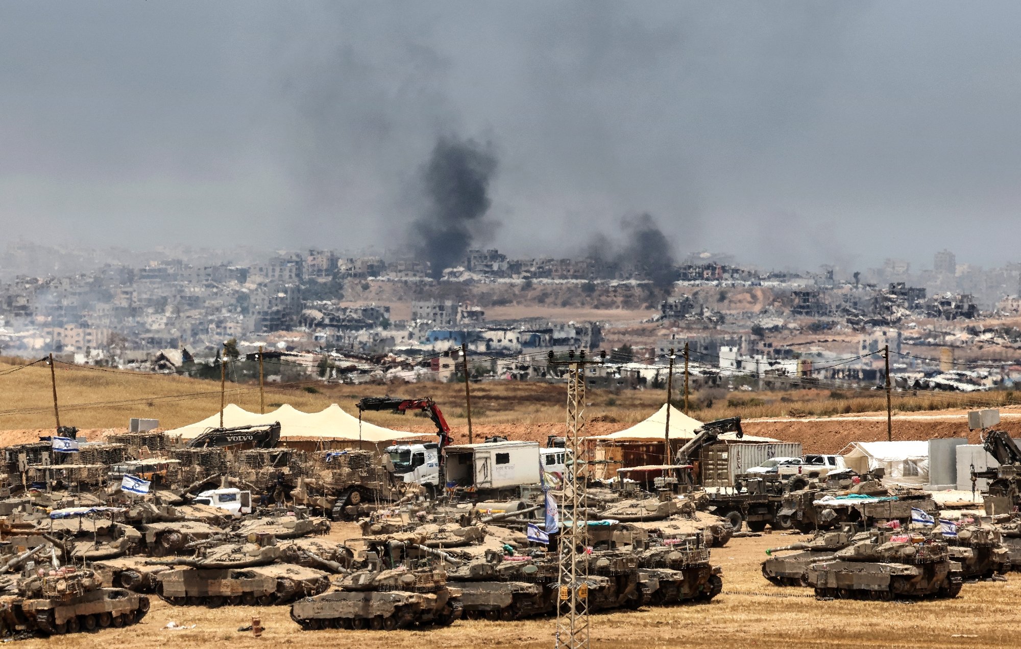 تصویر نشان‌دهنده یک منطقه جنگی با تانک‌ها و دود سیاه از انفجارها در دوردست است.