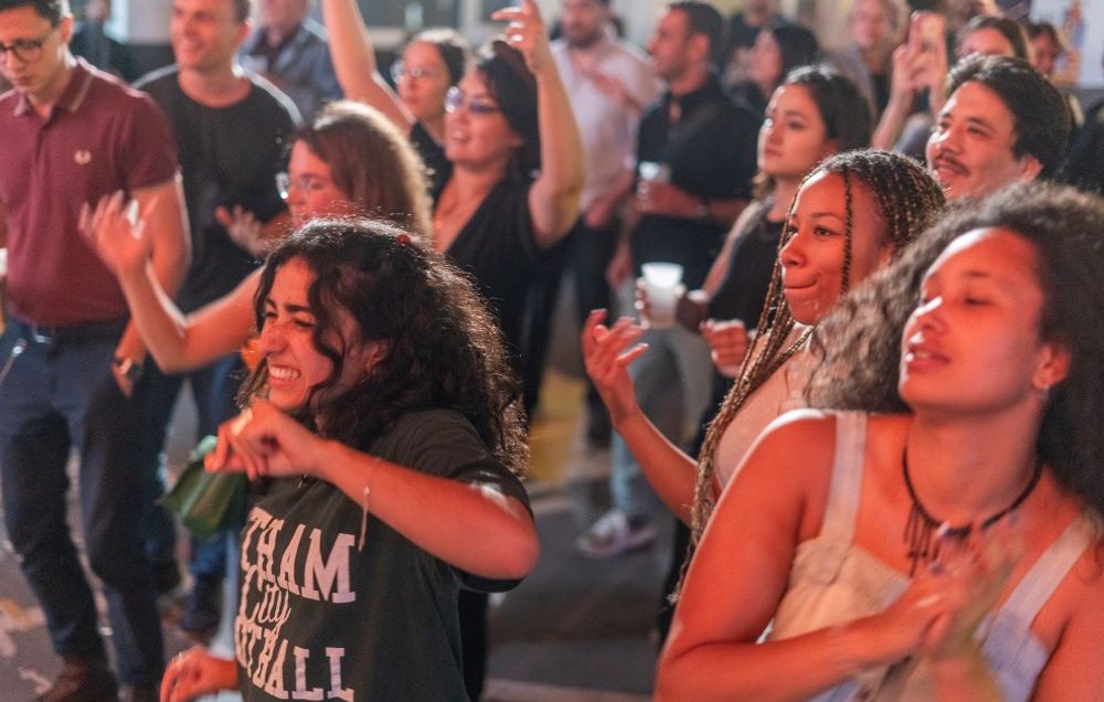 L'image montre un groupe de personnes en train de danser et de profiter d'une ambiance festive. On peut voir des personnes de différents âges, souriantes et s'amusant. Certaines gesticulent au rythme de la musique, tandis que d'autres sont concentrées sur le moment. L'éclairage est chaleureux, ce qui contribue à l'atmosphère joyeuse de l'événement.