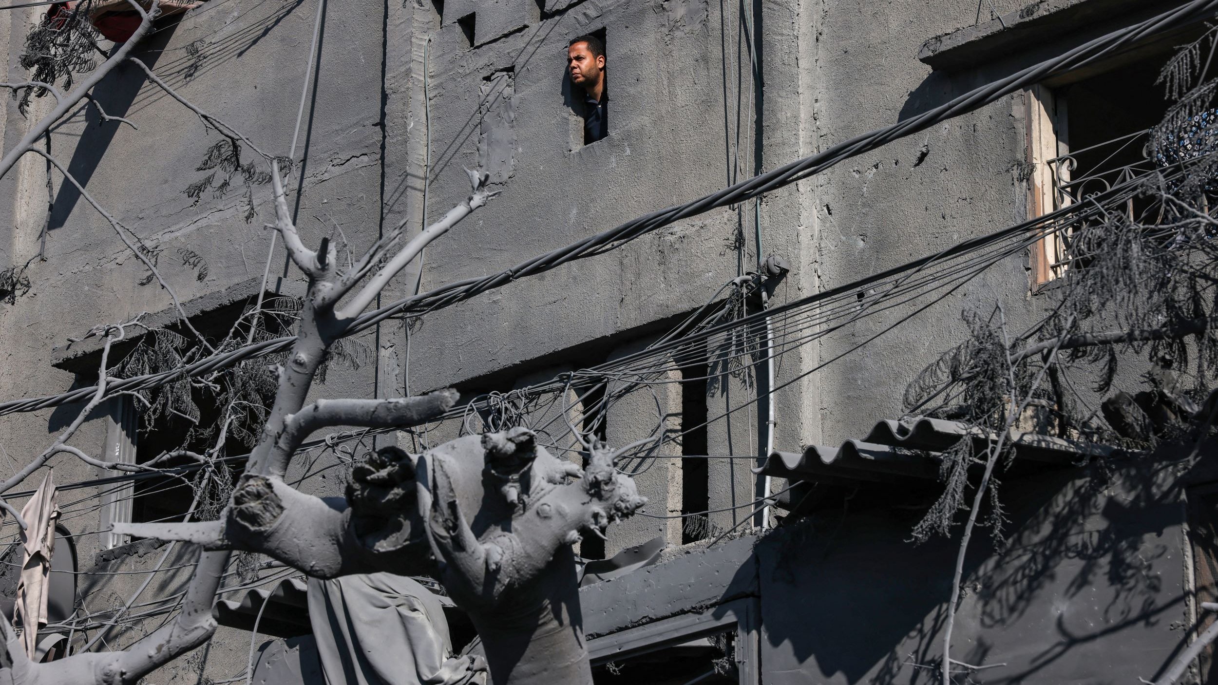 L'immagine mostra un edificio gravemente danneggiato, con i muri di cemento visibilmente rovinati. Da una finestra rotta, si intravede una persona che osserva l'esterno, mentre nel primo piano si trova una statua, anch'essa coperta di polvere e detriti. Le linee elettriche pendono disordinatamente, e l'atmosfera è cupa a causa dei colori grigi e della desolazione del contesto. La scena sembra suggerire un ambiente colpito da un evento catastrofico, con evidenti segni di distruzione e abbandono.