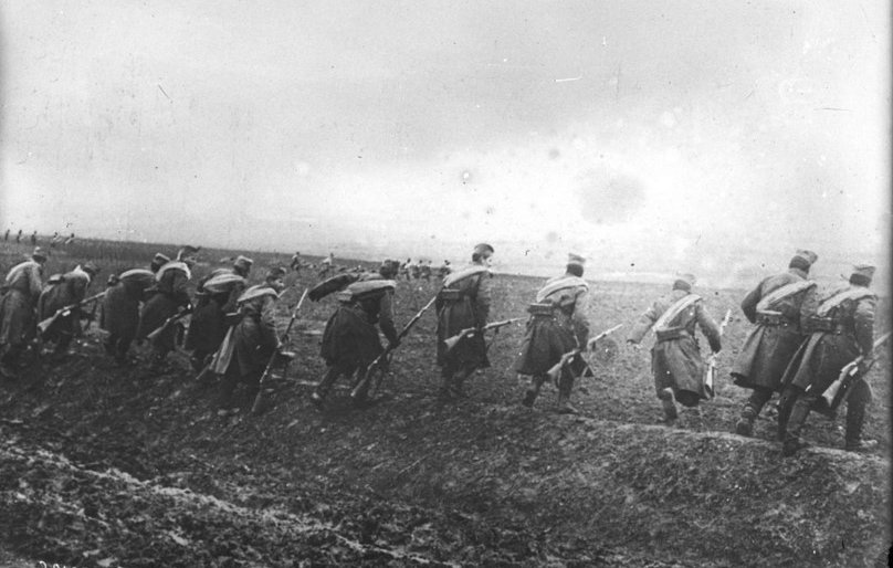 L'image montre un groupe de soldats marchant en formation sur un terrain accidenté. Ils portent des uniformes militaires, avec des chapeaux distinctifs, et semblent se déplacer dans une direction déterminée. Le paysage est marqué par un ciel nuageux et une ambiance sombre, suggérant peut-être un contexte de guerre ou de mouvement stratégique dans un environnement rural. Le sol est boueux, ce qui rend leur avance difficile. L'image est en noir et blanc, renforçant le caractère historique et poignant de la scène.