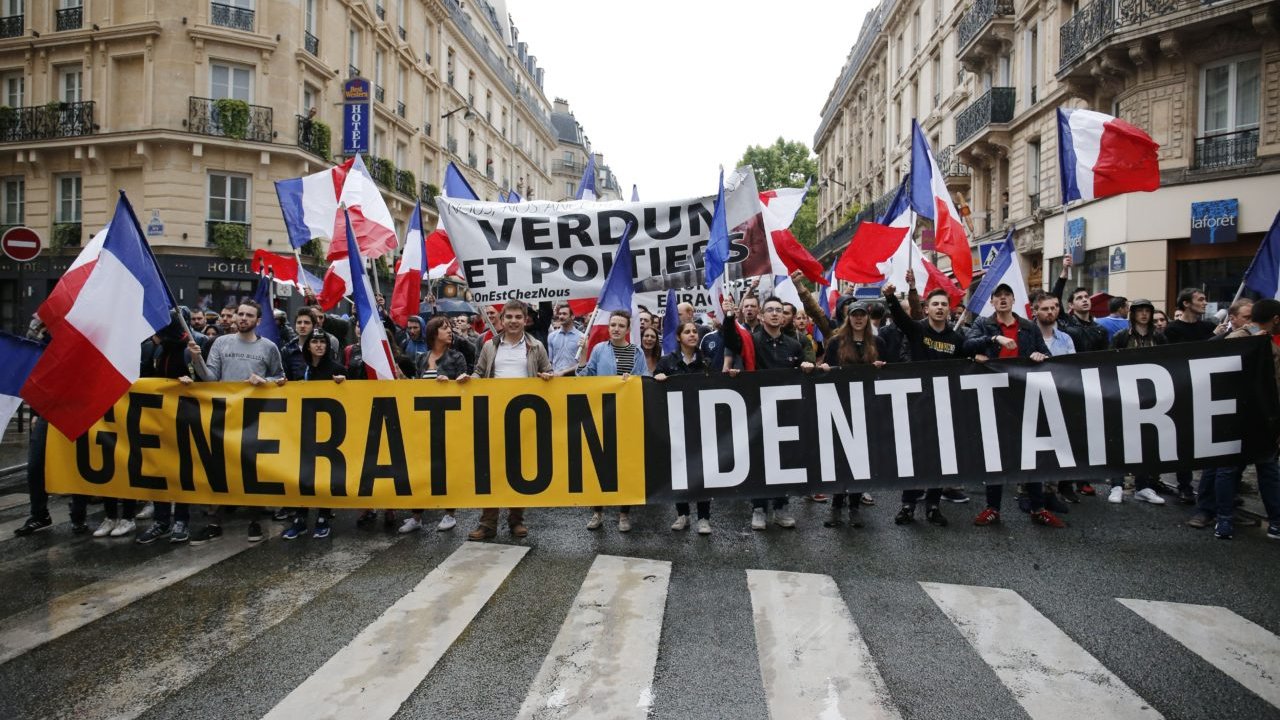L'image montre une manifestation dans une rue, où un groupe de personnes marche en brandissant des drapeaux français. Ils tiennent une grande banderole avec les mots "GENERATION IDENTITAIRE". En arrière-plan, d'autres banderoles sont visibles, affichant des slogans supplémentaires. L'atmosphère semble énergique et engagée, avec des participants portant des vêtements variés. Les bâtiments en arrière-plan reflètent un cadre urbain typique.