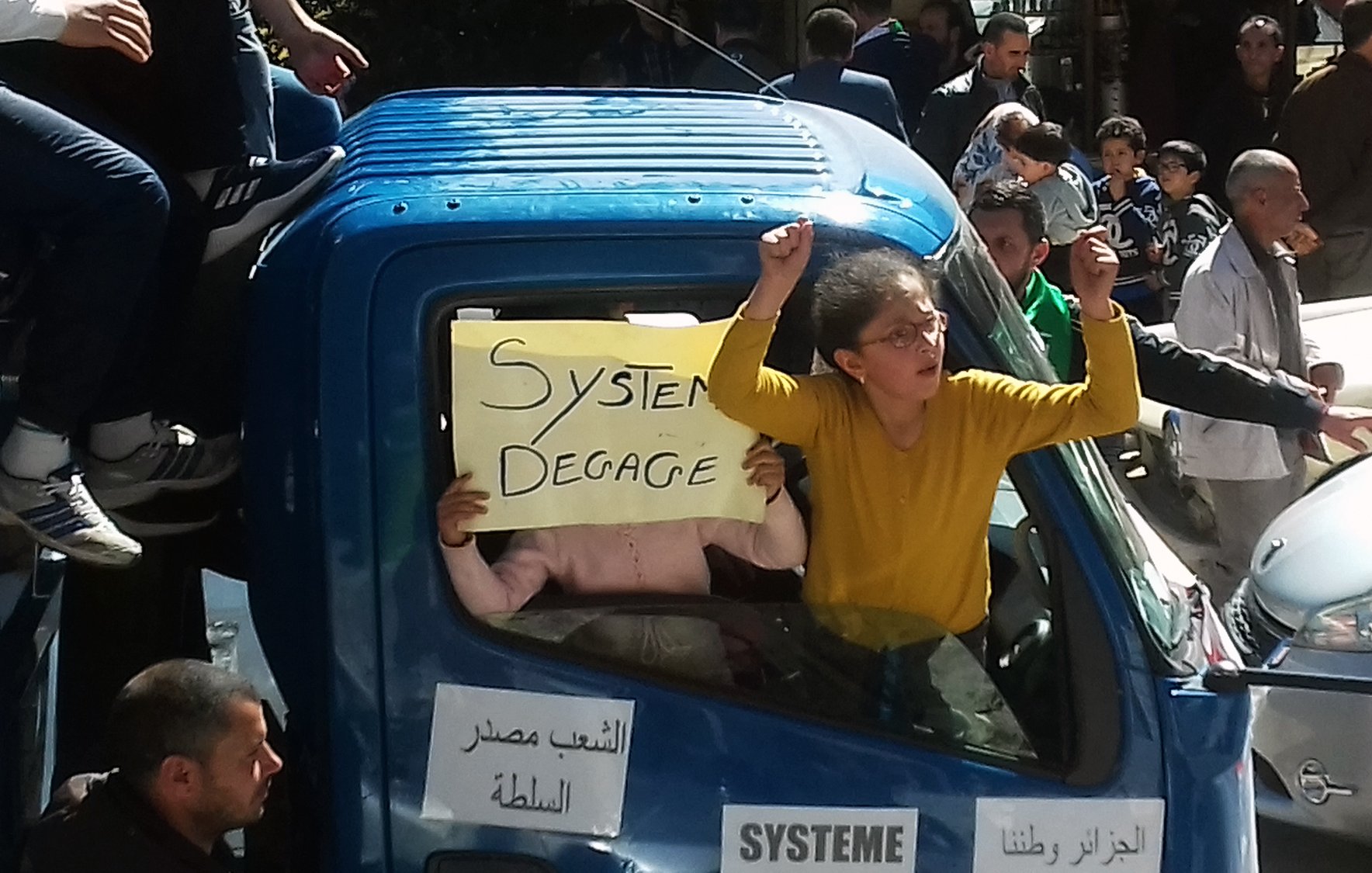 The image shows a group of people in a demonstration or protest, focused around a blue truck. One person is prominently displaying a sign that reads "Systeme Degage," indicating a call for the system to go or be removed. In the truck, another individual can be seen holding a sign. The atmosphere appears energetic, with participants expressing their views passionately. The surrounding crowd suggests a larger movement, likely addressing political or social issues.