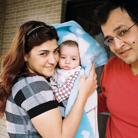 Sur cette image, on voit un couple et un bébé. La femme, à gauche, sourit et tient le bébé enveloppé dans une couverture, tandis qu'un homme, à droite, regarde la caméra en souriant également. Ils se tiennent tous les trois dans un environnement intérieur, probablement chez eux, avec un mur en briques en arrière-plan. L'atmosphère générale est chaleureuse et affectueuse.