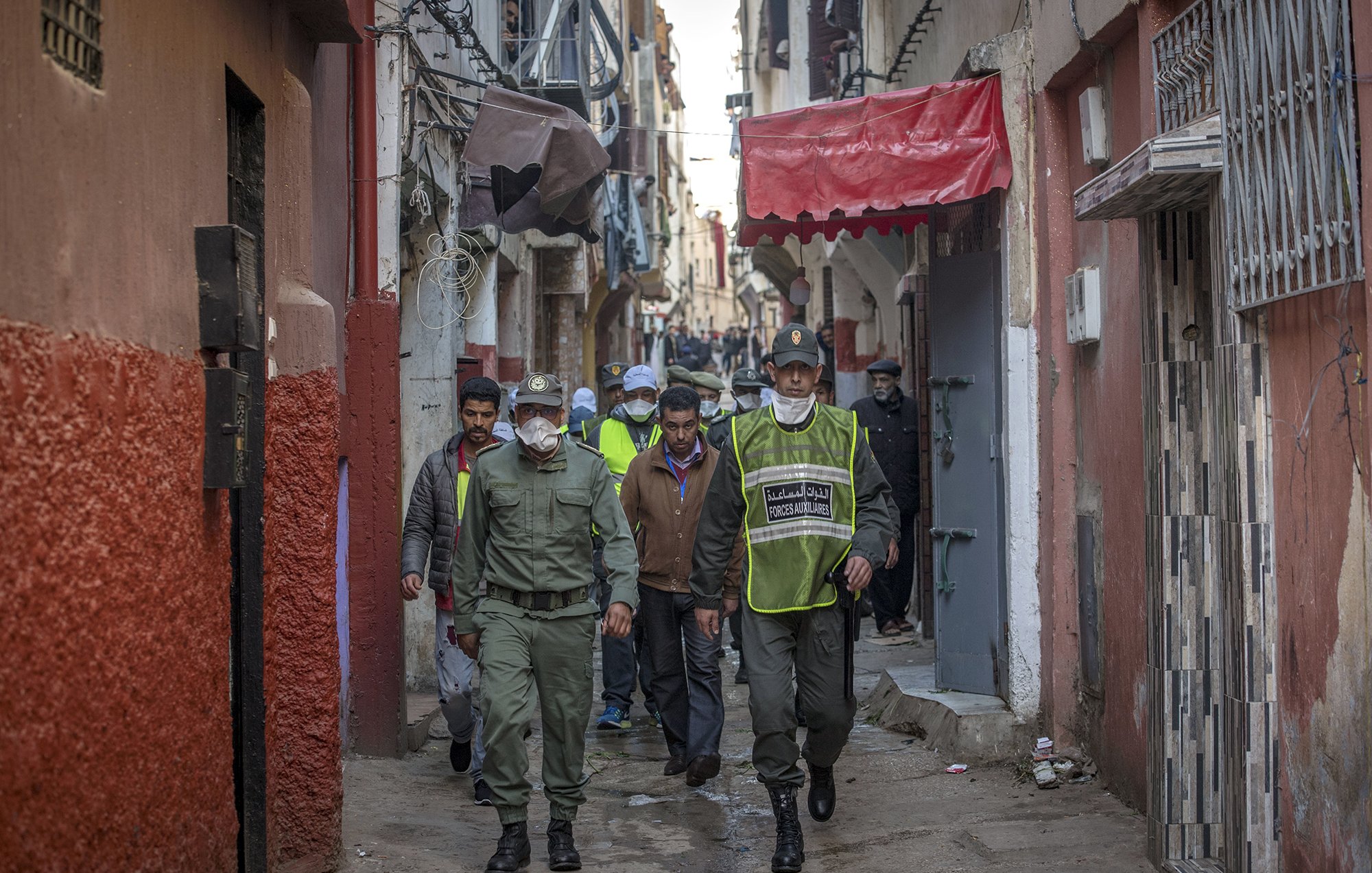 در این تصویر، یک کوچه باریک و قدیمی دیده می‌شود. در دو طرف کوچه، ساختمان‌ها با رنگ‌های مختلف نمایان هستند. گروهی از افرادی که لباس‌های فرم دارند، در حال قدم زدن در کوچه هستند. برخی از آن‌ها کلاه و ماسک به چهره دارند. محیط به نظر شلوغ و فعال می‌رسد و افراد مشغول به انجام کاری هستند. این تصویر فضایی محلی و اجتماعی را به نمایش می‌گذارد.