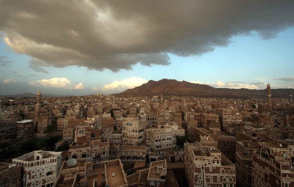 L'image montre une vue panoramique d'une ville traditionnelle, probablement située au Yémen, avec des bâtiments en adobe et des structures architecturales distinctives. On aperçoit des montagnes en arrière-plan, sous un ciel nuageux qui laisse entrevoir quelques éclaircies. La lumière du soleil couchant semble illuminer une partie de la ville, mettant en valeur les détails des constructions. L'ensemble dégage une atmosphère calme et pittoresque, reflétant la beauté du paysage urbain.