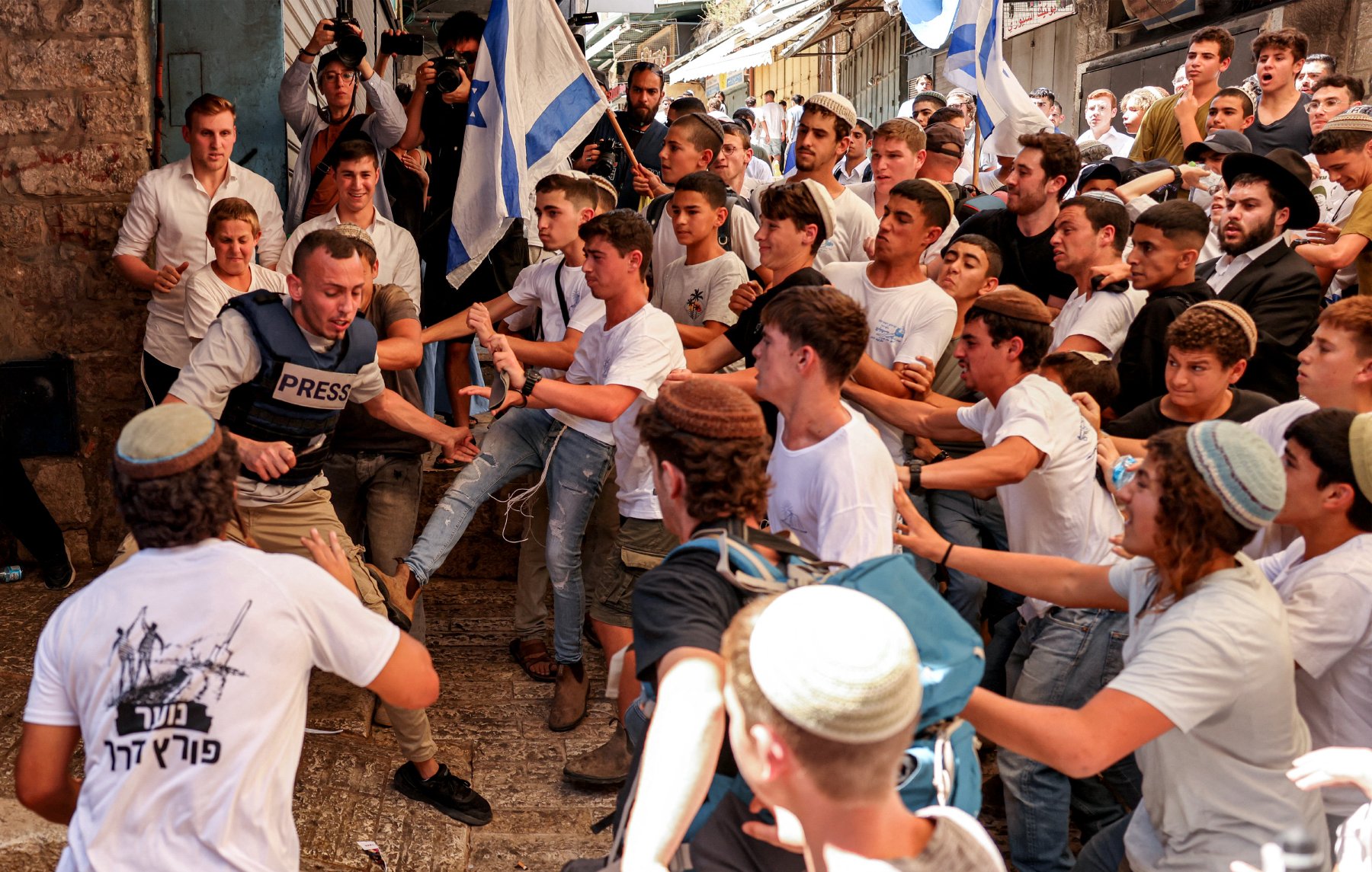 L'image montre un groupe animé de jeunes hommes dans une rue animée, probablement lors d'un événement ou d'une manifestation. Au premier plan, un homme portant un gilet "PRESS" semble être au centre de l'attention, entouré par plusieurs personnes qui cherchent à le repousser. Les participants portent des vêtements principalement blancs et bleu, suggérant une thématique nationale. L'atmosphère semble tendue, avec des expressions d'excitation et de conflit parmi la foule.