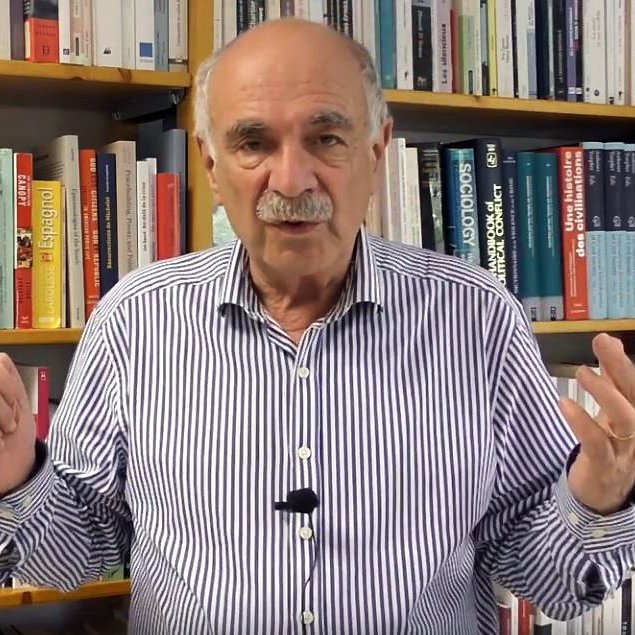 L'image montre un homme debout devant une étagère remplie de livres. Il porte une chemise à rayures et semble s'adresser à un public ou donner une présentation. Ses mains sont levées, ce qui indique qu'il est en train de faire un point ou d'expliquer quelque chose. L'arrière-plan est constitué de livres aux couvertures variées, suggérant un environnement intellectuel ou académique.