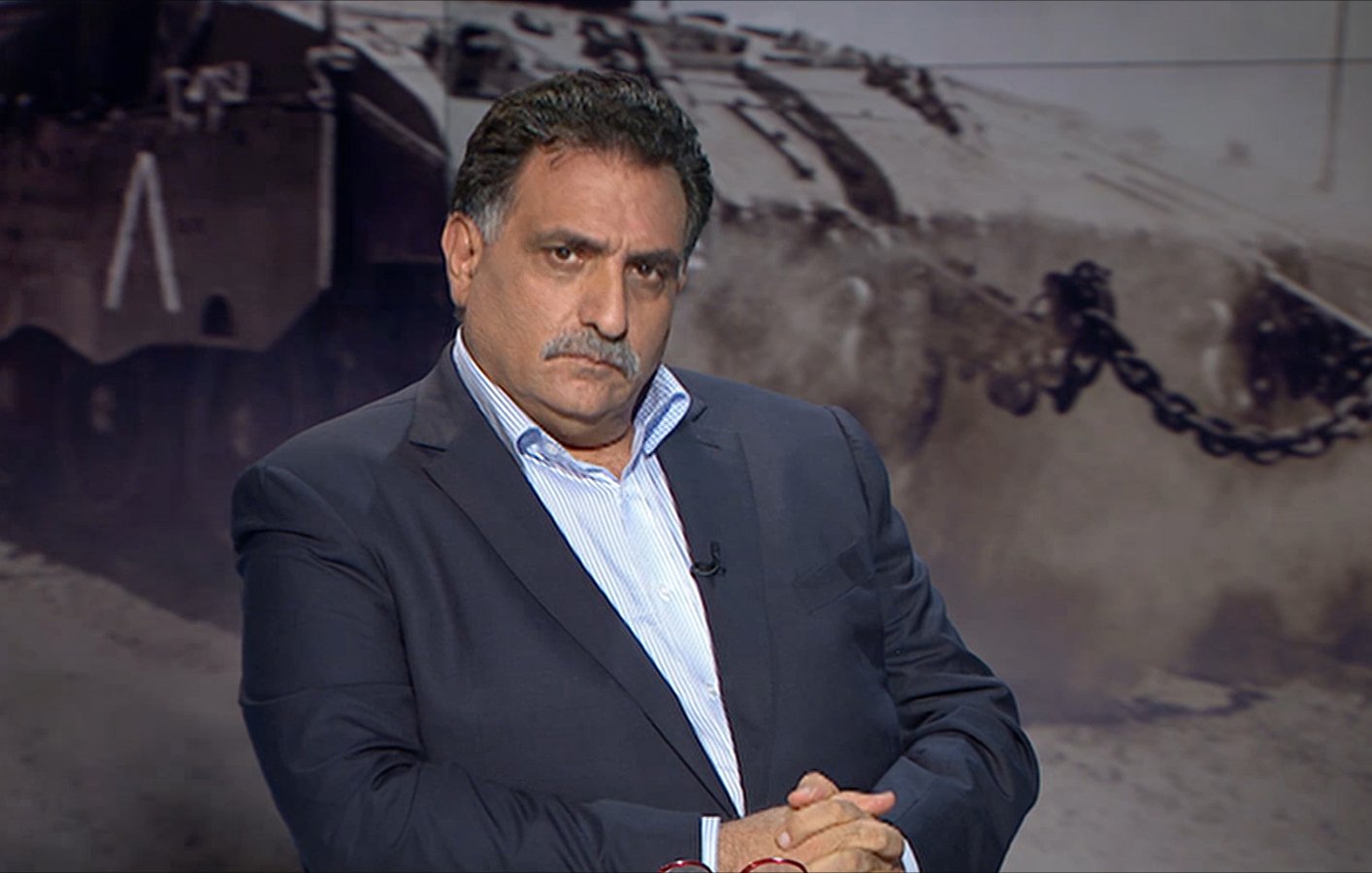 الصورة تظهر رجلًا يجلس بتعابير وجه جدية. يرتدي بدلة داكنة وقميص أبيض، ويداه متشابكتان أمامه. في الخلفية، يظهر مشهد غامض لمركبة عسكرية أو صورة حربية، مما يضيف جوًا من التوتر والدرامية للصورة. يبدو أن الرجل يتأمل أو يفكر بعمق.