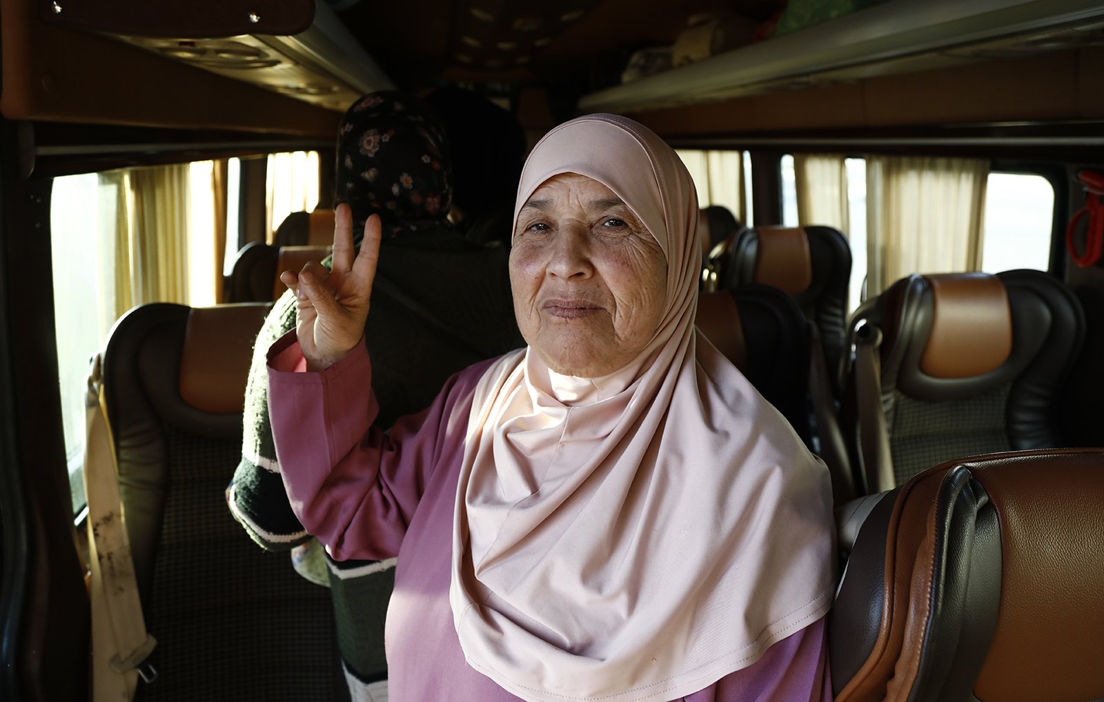L'image présente une femme âgée à l'intérieur d'un bus. Elle porte un hijab de couleur claire et un vêtement de style traditionnel. Elle fait un signe de paix avec ses doigts tout en souriant. En arrière-plan, on peut apercevoir d'autres passagers et l'intérieur du bus avec des sièges confortables. L'atmosphère semble chaleureuse et amicale.