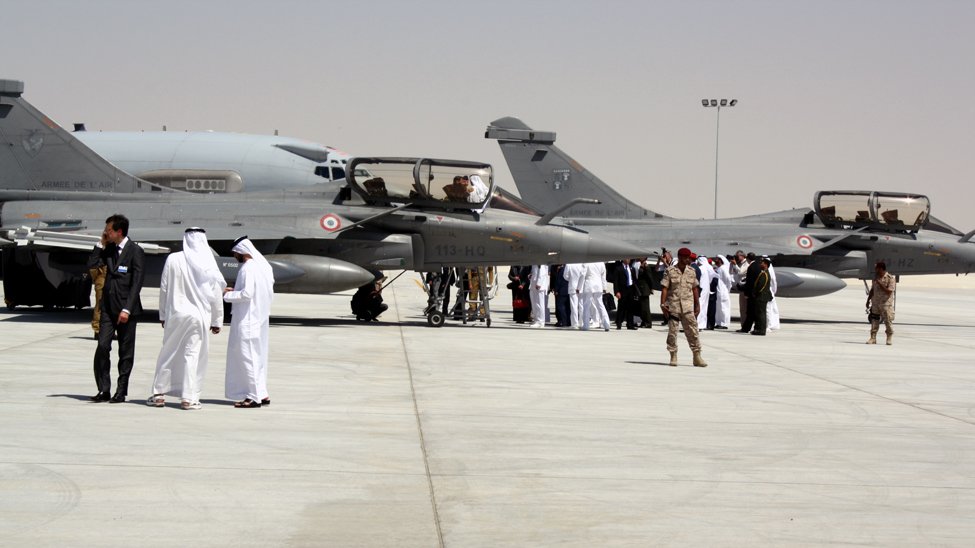 L'image montre une scène sur un tarmac d'aéroport militaire. On y voit plusieurs avions de chasse, probablement des Mirage ou des jets similaires, garés sur une surface bétonnée. Des membres d'équipage, habillés en uniforme, interagissent avec des personnes portant des costumes traditionnels. En arrière-plan, il y a d'autres personnes rassemblées, ce qui suggère un événement ou une cérémonie. Le cadre est désert, avec un ciel clair, ce qui indique qu'il pourrait s'agir d'une région aride.