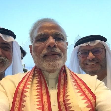L'image montre un groupe de trois hommes prenant un selfie, probablement dans un cadre officiel ou lors d'un événement. L'homme au centre porte un vêtement traditionnel avec un foulard coloré autour du cou. Les deux autres hommes à côté de lui portent des vêtements traditionnels du Moyen-Orient, incluant un keffieh et un costume, ajoutant à l'atmosphère culturelle de la scène. En arrière-plan, on aperçoit des bâtiments qui semblent être emblématiques, bien que les détails ne soient pas clairs. L'expression des visages est souriante et conviviale, suggérant une atmosphère amicale.