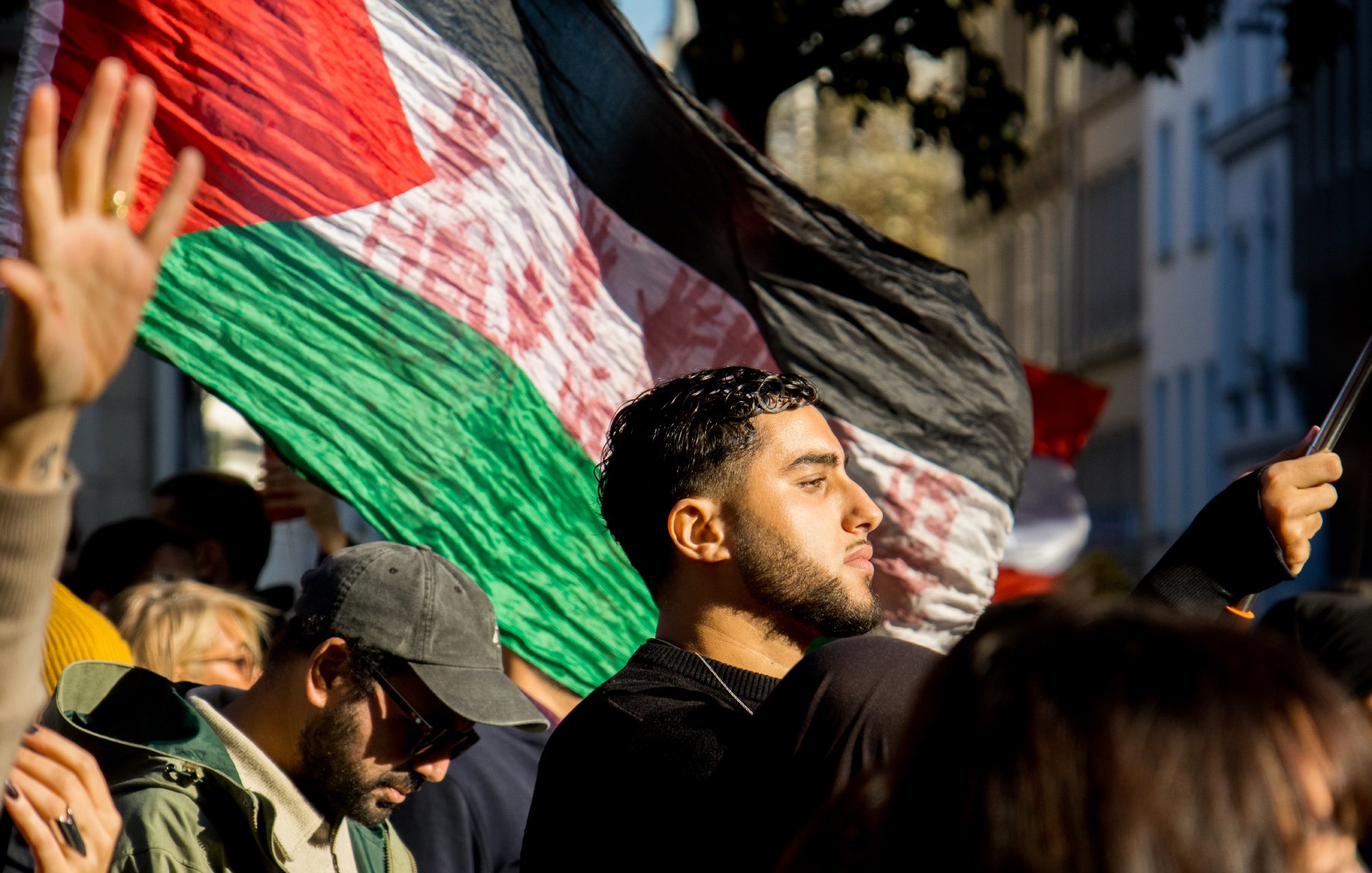 L'image montre une foule lors d'une manifestation. On peut voir une personne au premier plan tenant un grand drapeau aux couleurs du pays. Il y a également d'autres manifestants en arrière-plan, dont certains portent des lunettes de soleil et d'autres ont des expressions engagées. La lumière du soleil semble créer une ambiance dynamique et engagée, accentuant l'émotion de l'événement.