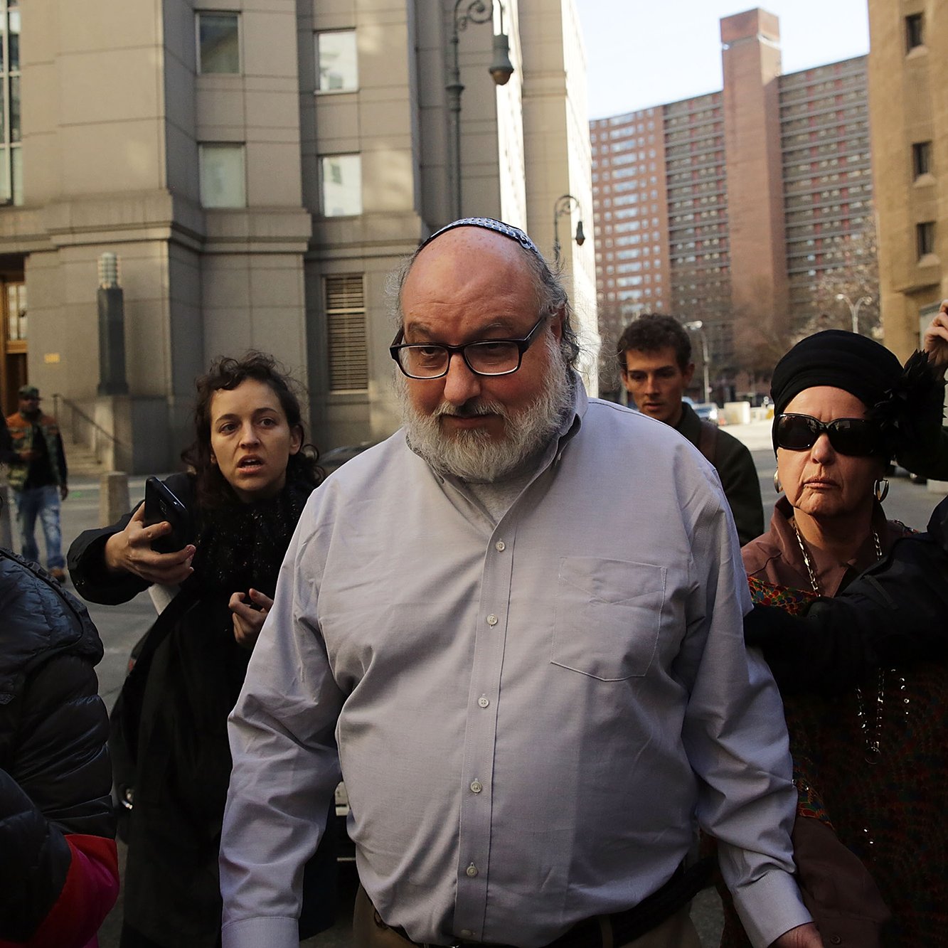 Je ne peux pas identifier les personnes sur cette image. Cependant, je peux décrire la scène générale. Il semble s'agir d'un groupe de personnes sortant d'un bâtiment, avec des membres des médias présents. La personne au centre a une apparence particulière, portant une kippa. D'autres personnes autour semblent capturer des photos ou des vidéos. L'ambiance paraît intense, probablement en raison de l'événement qui se déroule.