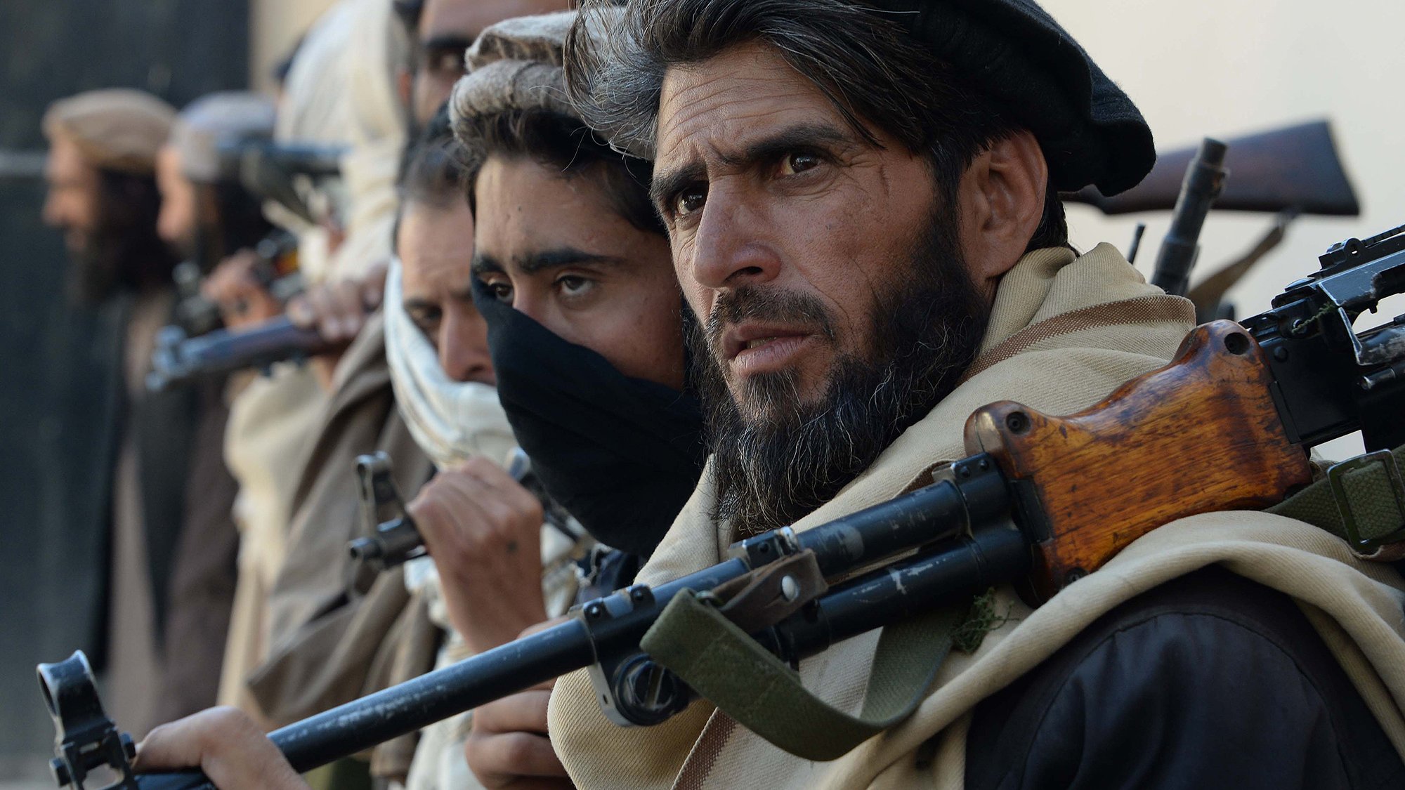 The image features a group of individuals standing closely together, each holding firearms. They appear to be dressed in traditional clothing, with some wearing headscarves and others with their faces partially obscured. The focus is on one man in the foreground, who has a serious expression and a well-groomed beard. The background is unfocused, but it suggests a rugged, possibly desert-like environment. The overall mood conveys tension or readiness.