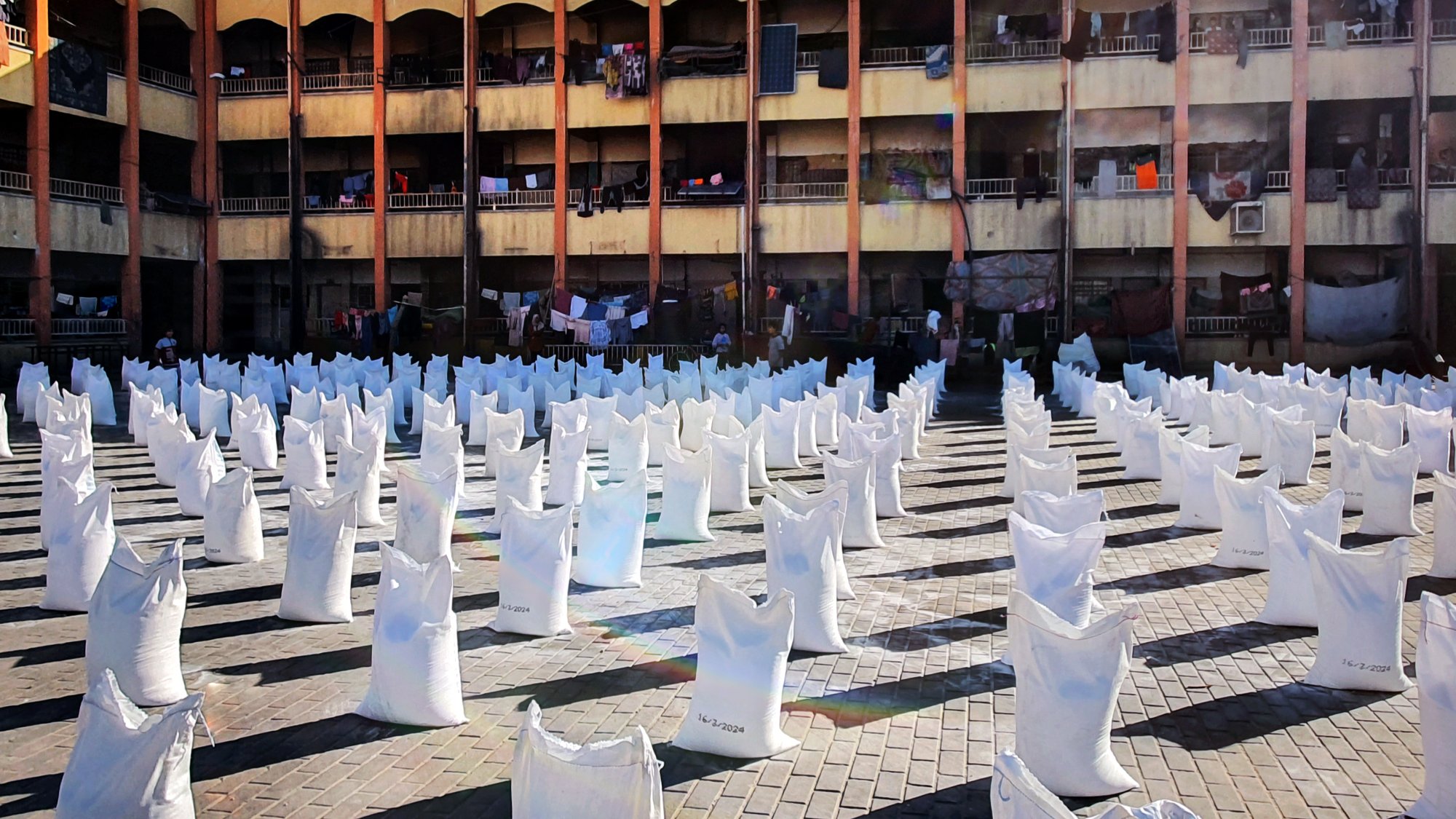 L'immagine mostra un'area all'aperto con molti sacchi bianchi disposti in file ordinate. Sullo sfondo si vede un edificio di più piani, con diversi balconi. Alcuni panni sono appesi ad asciugare tra i balconi. La scena è illuminata dalla luce del sole, creando un'atmosfera vivace e attiva. Il pavimento è costituito da mattonelle in cui si possono notare i segni del tempo. L'insieme suggerisce un contesto urbano con attività legate alla distribuzione di beni.