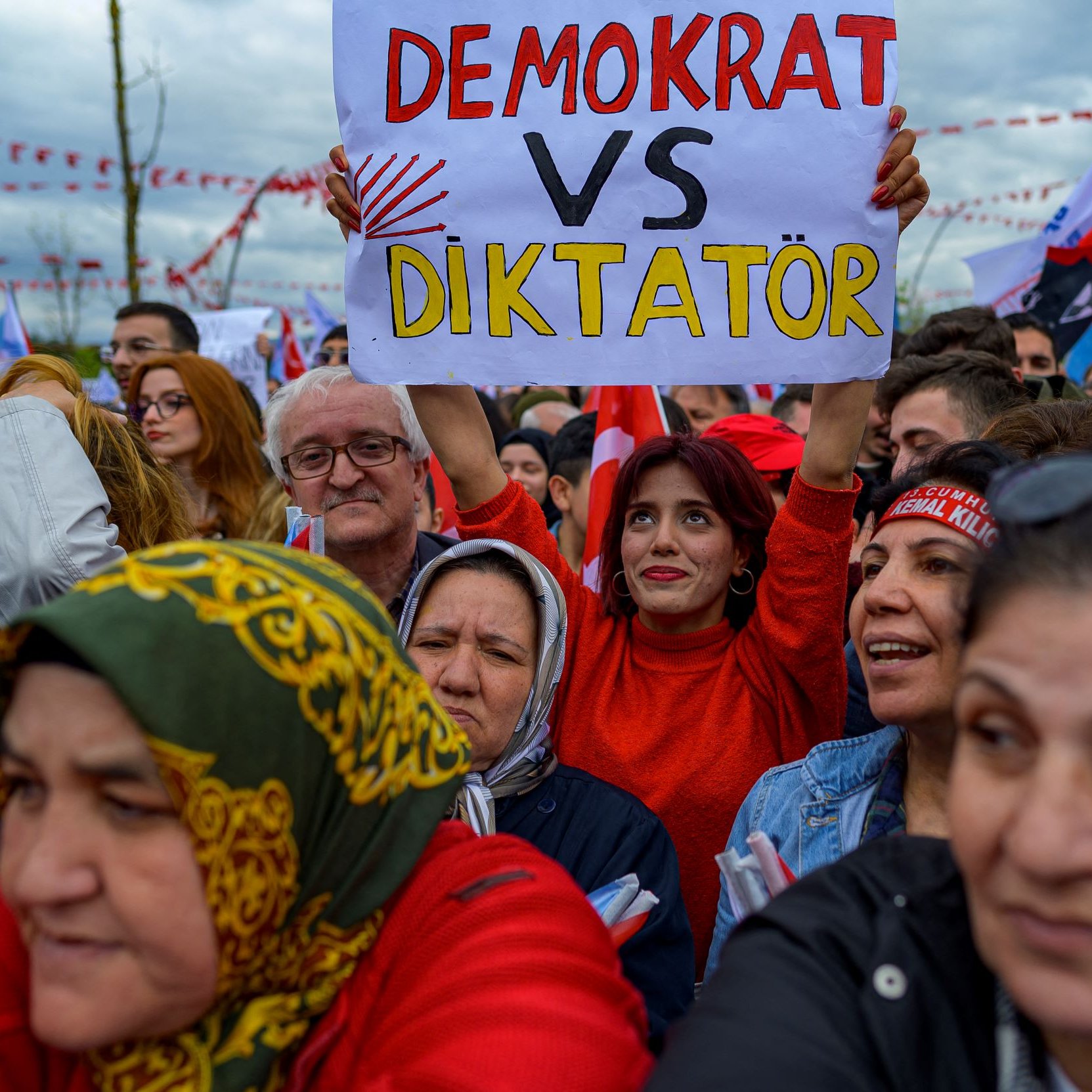 L'image montre une foule rassemblée lors d'un événement, probablement une manifestation ou un rassemblement politique. Au premier plan, on voit plusieurs personnes, dont une femme tenant une pancarte sur laquelle est inscrit "DEMOCRAT VS DIKTATÖR". Les participants semblent engagés et déterminés, et les expressions sur leurs visages reflètent des émotions fortes. On peut également observer des drapeaux et des banderoles en arrière-plan, suggérant un contexte de mobilisation politique.