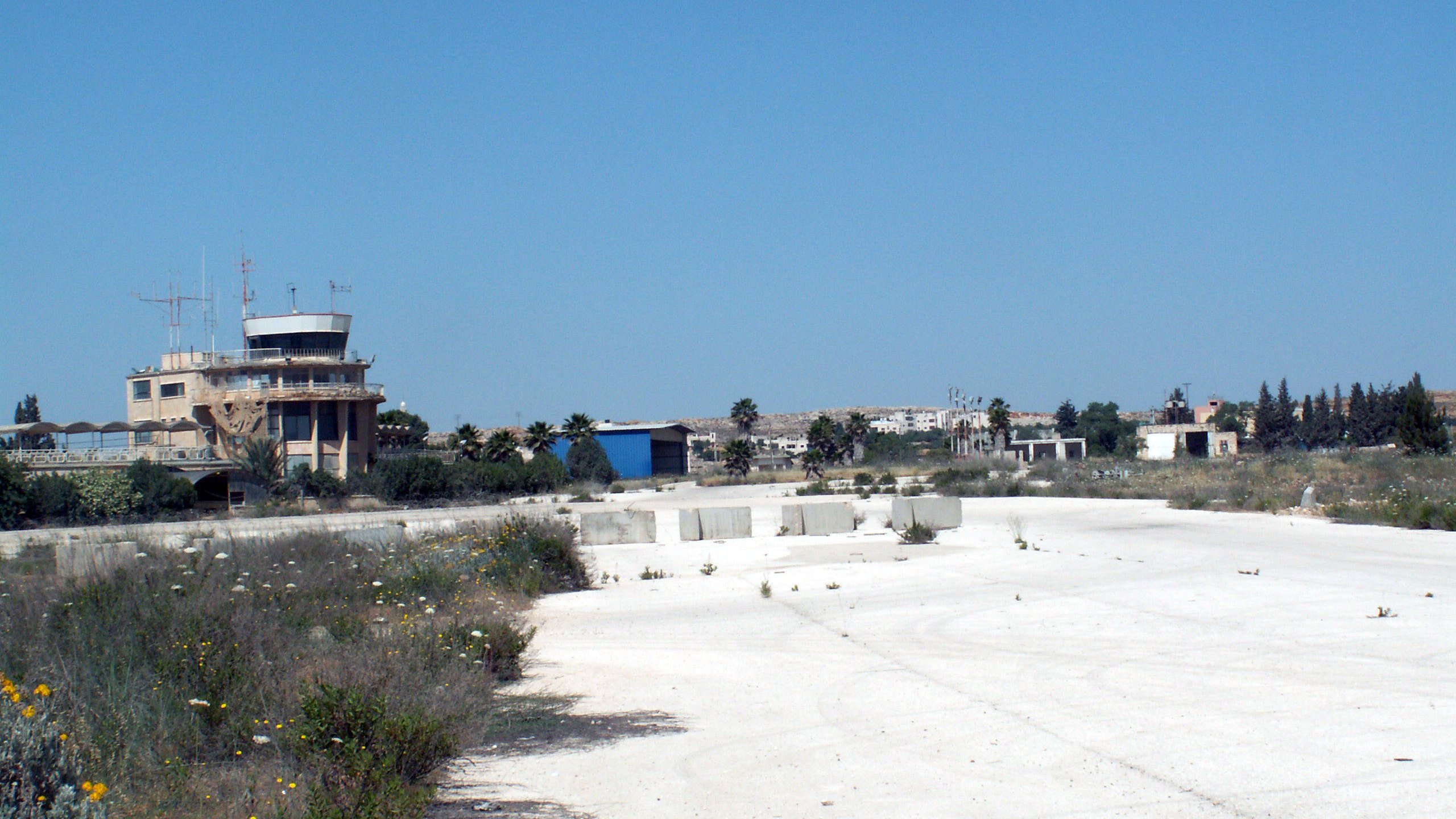 La imagen muestra un paisaje de un área desierta, posiblemente un antiguo aeropuerto o base aérea. En el fondo se observa una torre de control o edificio administrativo, rodeado de vegetación y estructuras en desuso. El suelo es de color claro y parece estar cubierto de hierbas y flores silvestres, lo que sugiere abandono. El cielo es de un azul despejado, lo que le da un contraste al paisaje.