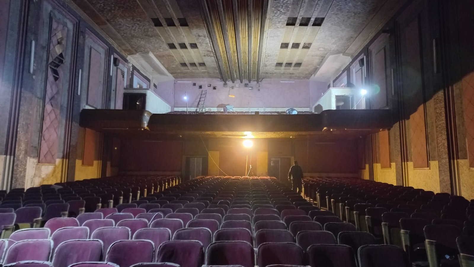 L'image montre l'intérieur d'un théâtre ancien. On peut voir des rangées de sièges en velours, qui semblent un peu usés, dirigées vers une scène surélevée. La scène est éclairée par une lumière vive, tandis que le reste de la salle est dans une ambiance plus sombre. On aperçoit également des détails architecturaux au plafond et sur les murs, qui évoquent un style vintage. Un homme se tient debout au fond, ajoutant une touche humaine à cet espace, qui dégage une atmosphère à la fois nostalgique et mystérieuse.