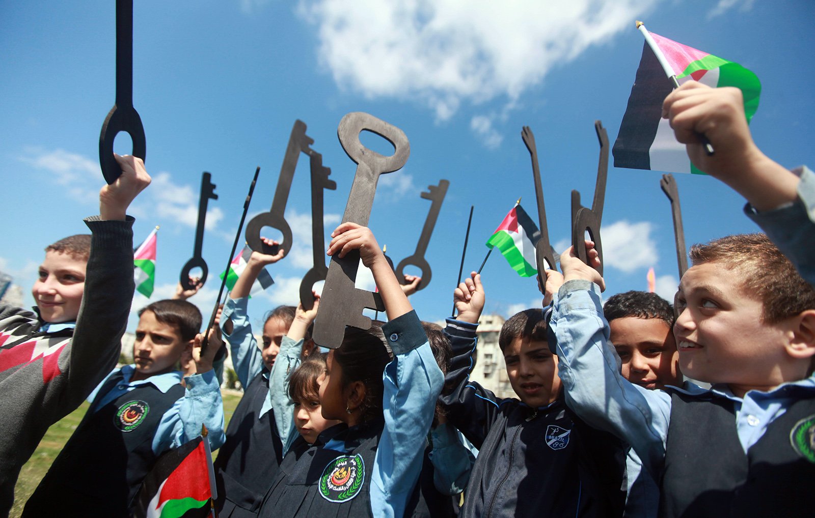 الصورة تظهر مجموعة من الأطفال يقفون في الهواء الطلق، يحملون مفاتيح كبيرة وأعلام فلسطينية. يبدو أنهم يعبرون عن تأييد لقضية معينة أو احتفال بذكرى تاريخية. الأطفال مبتسمون ومتحمسون، مما يعكس روح الفرح والاحتفاء والوعي بالقضايا المتعلقة بفلسطين. السماء صافية وملونة بالغيوم، مما يعطي شعوراً بالحيوية والنشاط.