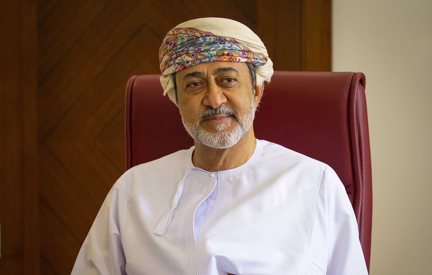 الصورة تظهر شخصًا يجلس على كرسي. يرتدي الملابس التقليدية العمانية، حيث يظهر بجلابية بيضاء وعصابة رأس مزخرفة. في يده يوجد خنجر عماني تقليدي. خلفه خلفية خشبية بسيطة. يعكس تعبير الوجه الهدوء والثقة.