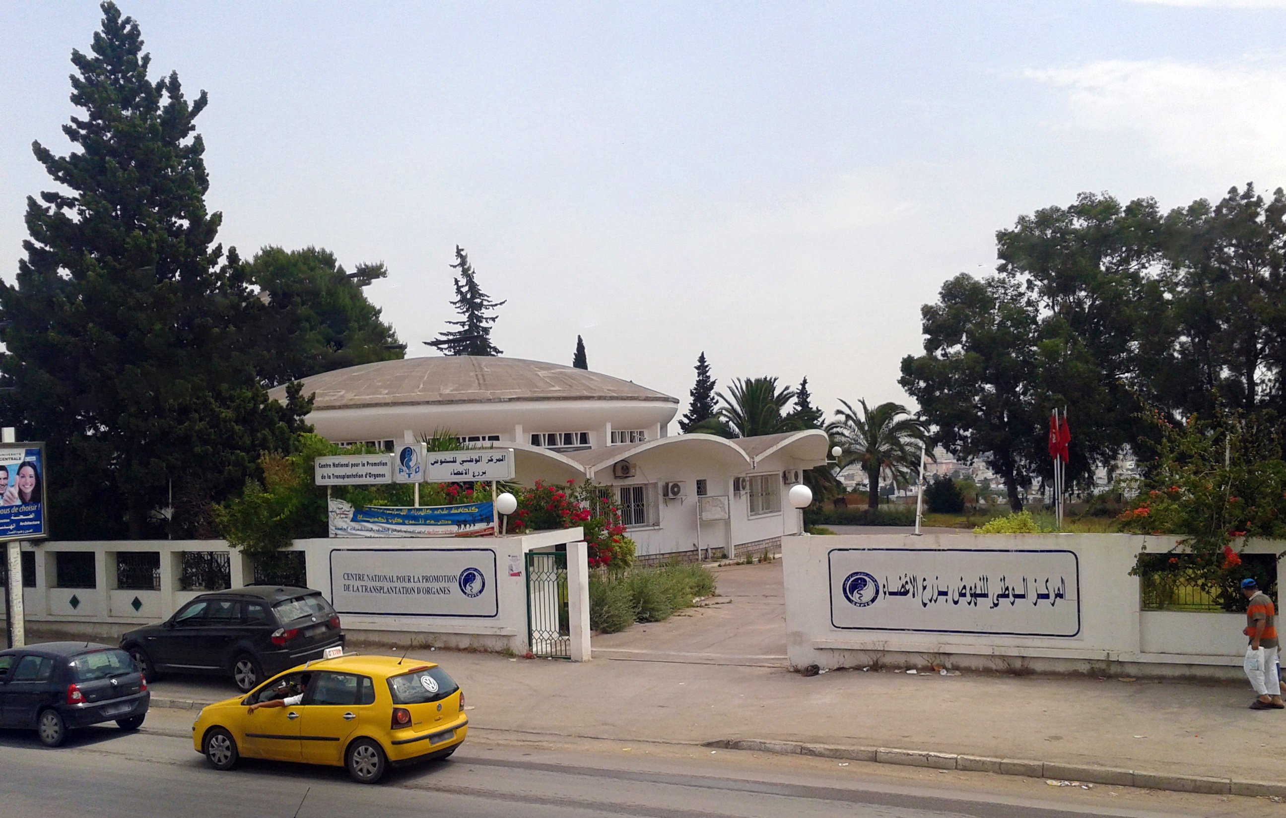 The image depicts a modern building with a dome-shaped roof, likely serving as a public institution or center. The architecture features a combination of curved and angular elements, with large windows that allow natural light inside. Surrounding the building are trees and greenery, contributing to a pleasant atmosphere. In front of the building, there are vehicles parked, including a yellow taxi, indicating it is situated near a road. There are also banners displaying text, possibly providing information about the institution. A flag can be seen on a pole, suggesting this might be a governmental or community facility. Overall, the scene conveys a sense of public service in a well-maintained environment.