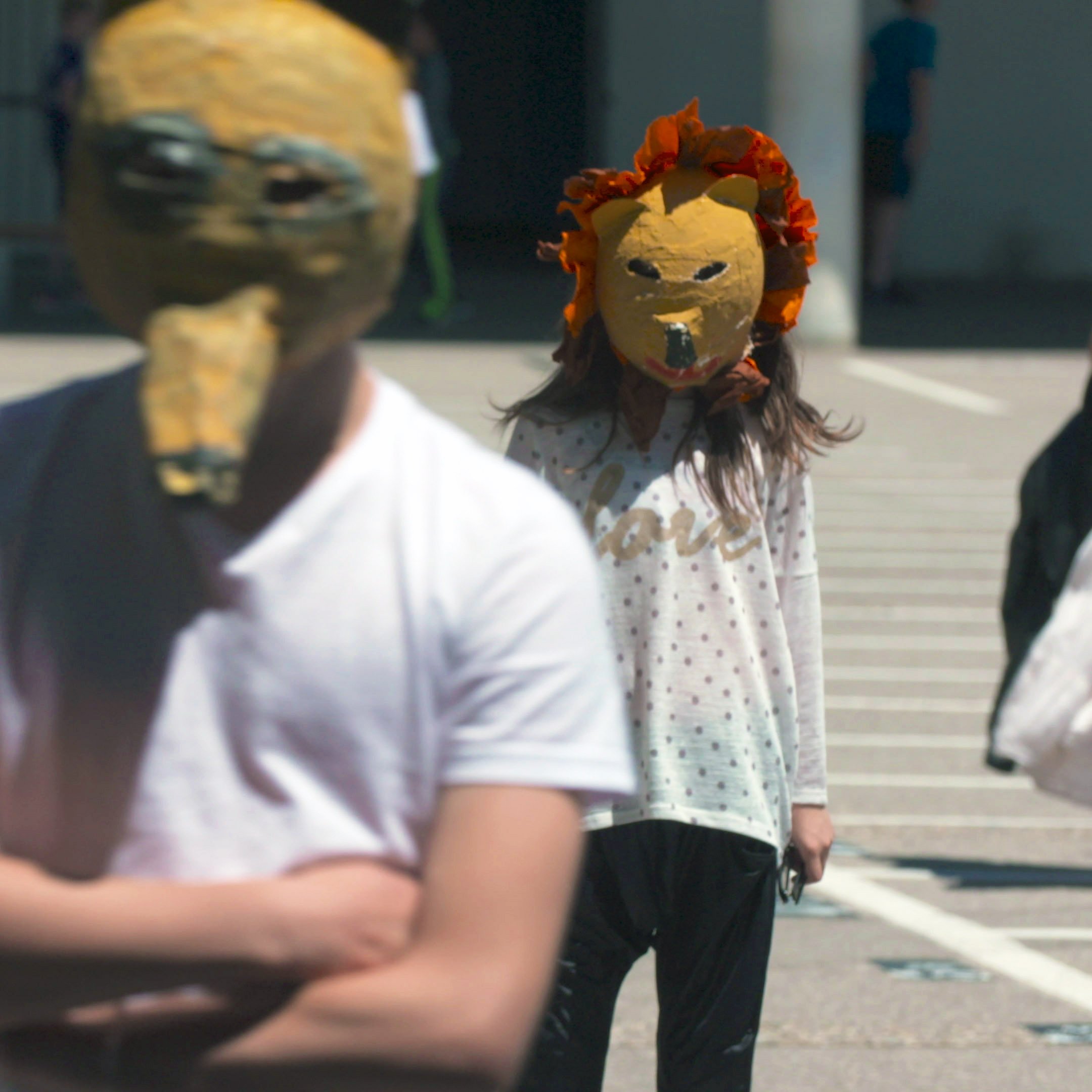 L'image montre deux personnes portant des masques originaux. L'un des masques a une forme de tête d'animal, tandis que l'autre est décoré de fleurs, évoquant un lion. Les personnes semblent se promener dans un espace extérieur, peut-être une place ou une cour. L'environnement est lumineux et rappelle une scène de performance artistique ou d'un événement communautaire.