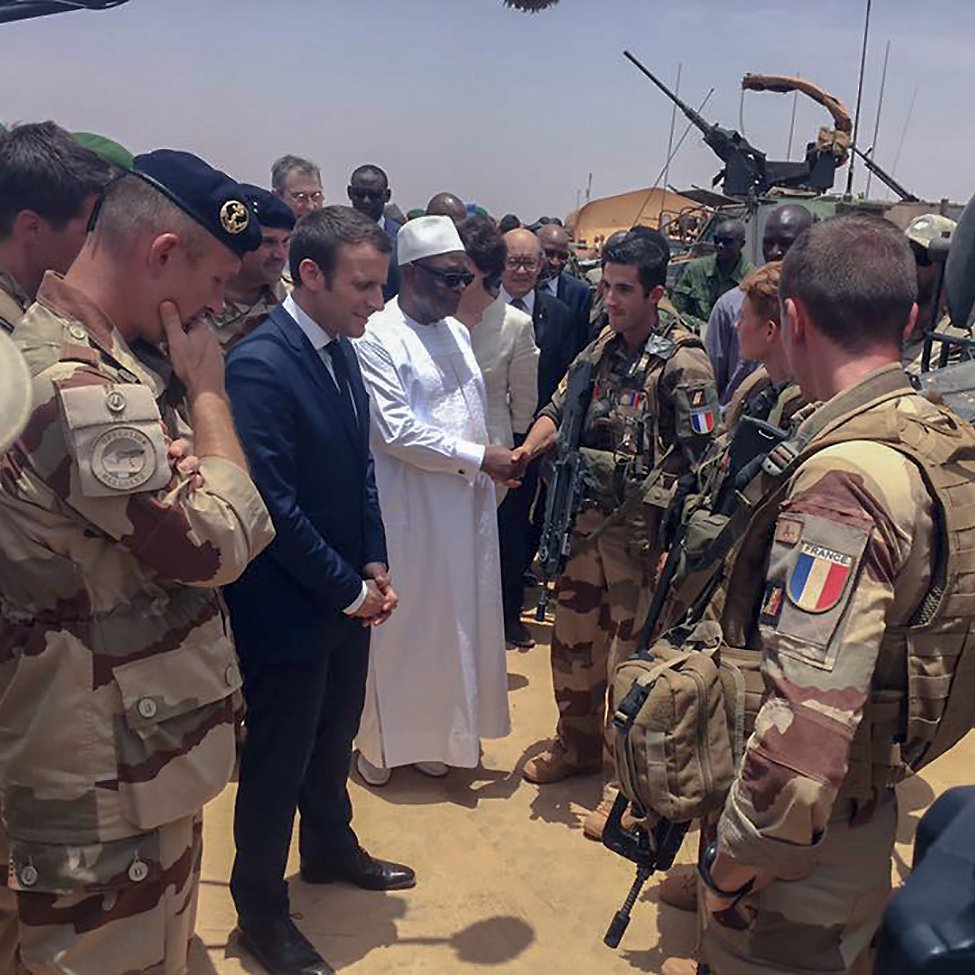 L'image montre un groupe de militaires et de responsables, probablement en situation de rencontre officielle dans un environnement désertique. Au centre, on peut voir un homme en costume, représentant vraisemblablement un leader, en train de discuter avec des soldats équipés de matériel militaire. Les militaires sont en uniforme et semblent engagés dans une mission, tandis que d'autres personnes obéissent à la dynamique de la réunion. L'atmosphère paraît sérieuse et solennelle.
