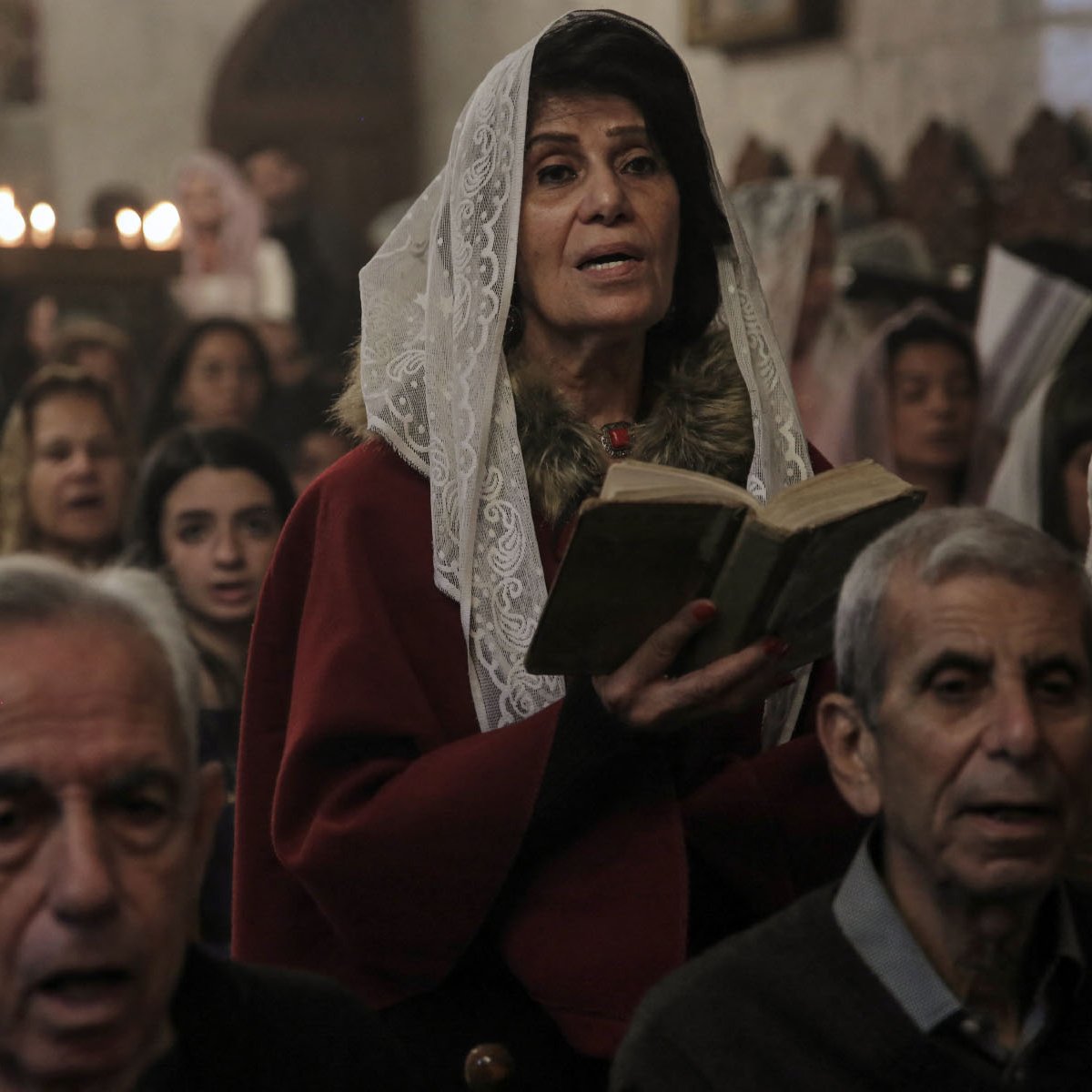 L'image montre un groupe de personnes rassemblées dans un lieu de culte. Au premier plan, une femme portant un vêtement rouge et un voile lit un livre, probablement un texte religieux. En arrière-plan, d'autres personnes, dont certaines semblent chanter ou prier, sont visibles. L'atmosphère semble solennelle, éclairée par des bougies qui créent une ambiance chaleureuse. Les visages des participants expriment une concentration et une dévotion.