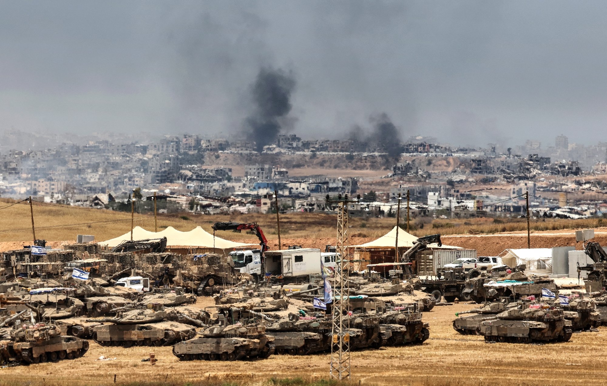 Terreno desolato con carri armati, fumi di incendi e un paesaggio urbano distrutto sullo sfondo.