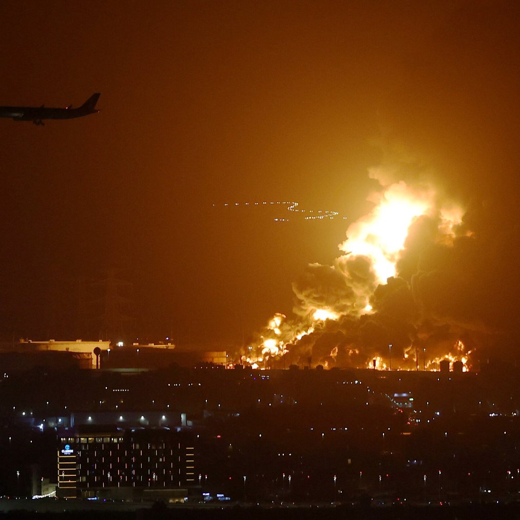 L'image montre un paysage nocturne éclairé par des flammes intenses, suggérant un incendie ou une explosion. On peut voir des colonnes de feu se levant dans le ciel, créant une ambiance dramatique. À l'arrière-plan, un avion est en train de survoler la scène, ce qui indique la proximité d'un aéroport. En bas, des lumières de la ville sont visibles, ajoutant un contraste entre l'urbanité et la catastrophe en cours.