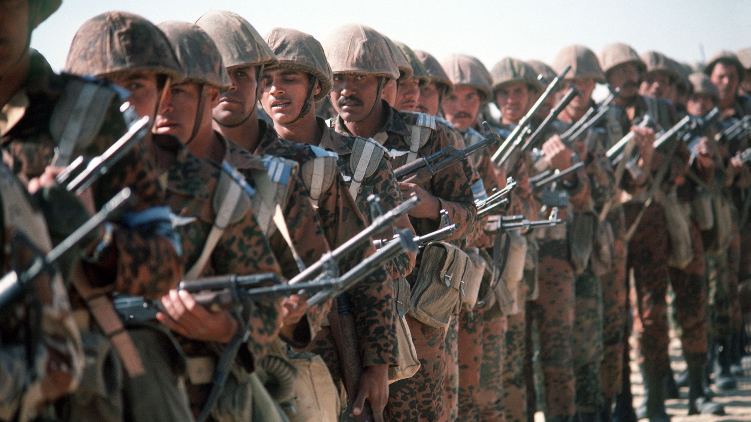 L'image montre une formation de soldats en tenue militaire, portant des casques et armés de fusils. Ils sont alignés en rangée, faisant face à l'objectif de la caméra. Leurs uniformes sont camouflés, et les expressions de leurs visages reflètent la concentration et la détermination. L'arrière-plan est désertique, suggérant un environnement de préparation militaire.