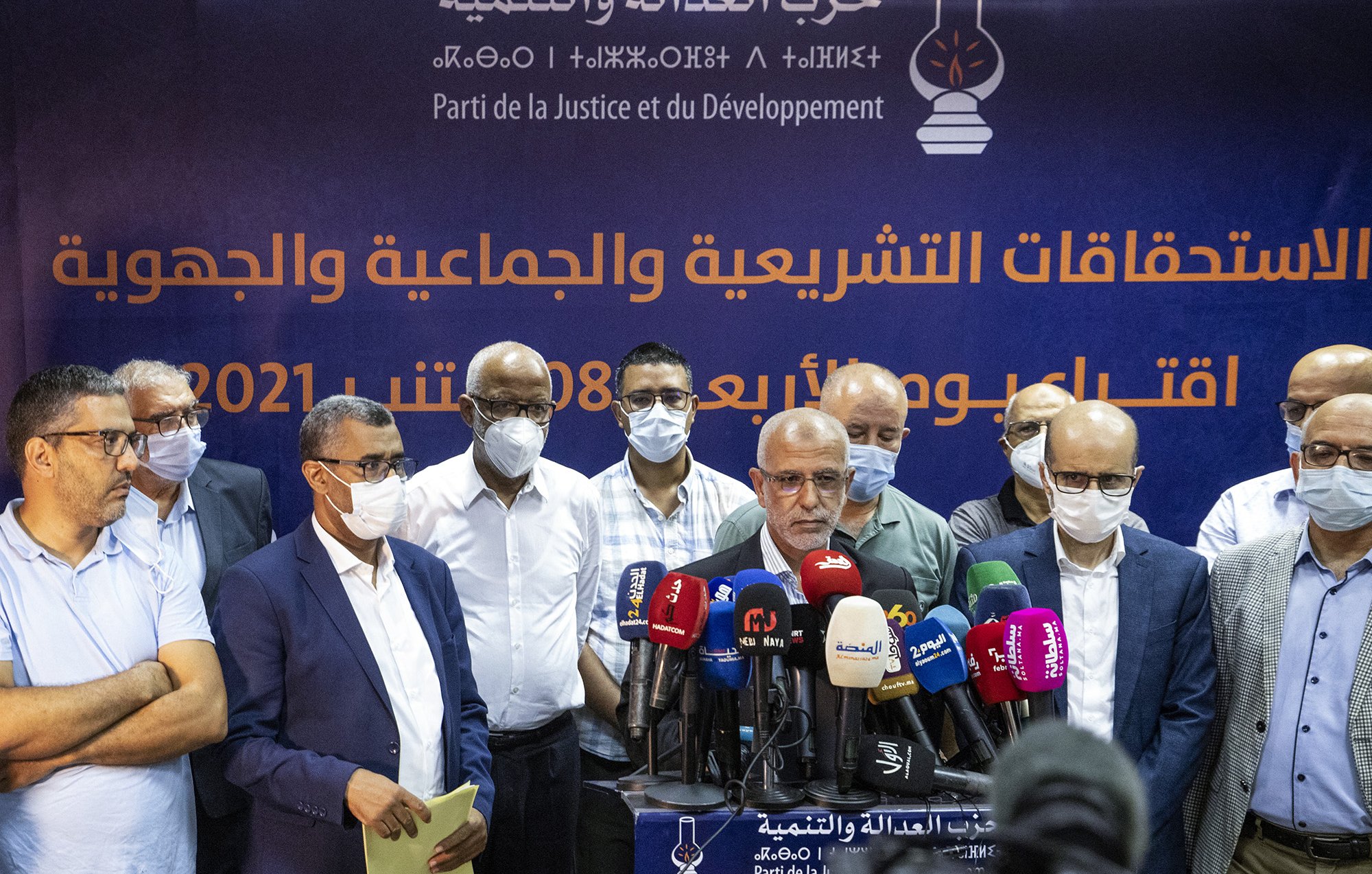 Sur l'image, un groupe d'hommes se tient devant une bannière affichant le nom d'un parti politique, le Parti de la Justice et du Développement. Les hommes portent des masques de protection, ce qui suggère un contexte de santé publique. Ils semblent s'apprêter à faire une déclaration ou une conférence de presse, entourés de microphones et de caméras. L'atmosphère semble sérieuse et professionnelle.