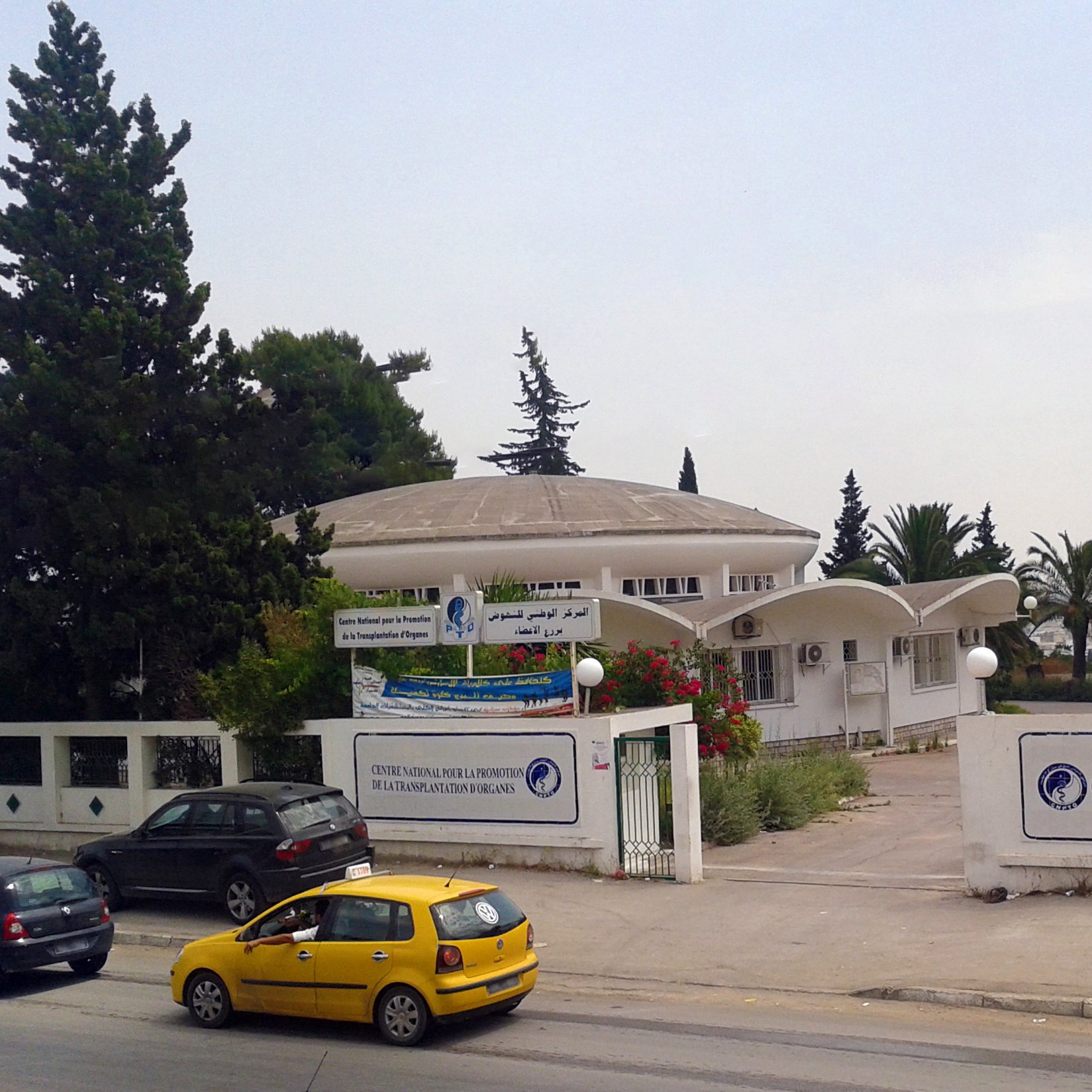 The image depicts a modern building with a dome-shaped roof, likely serving as a public institution or center. The architecture features a combination of curved and angular elements, with large windows that allow natural light inside. Surrounding the building are trees and greenery, contributing to a pleasant atmosphere. In front of the building, there are vehicles parked, including a yellow taxi, indicating it is situated near a road. There are also banners displaying text, possibly providing information about the institution. A flag can be seen on a pole, suggesting this might be a governmental or community facility. Overall, the scene conveys a sense of public service in a well-maintained environment.