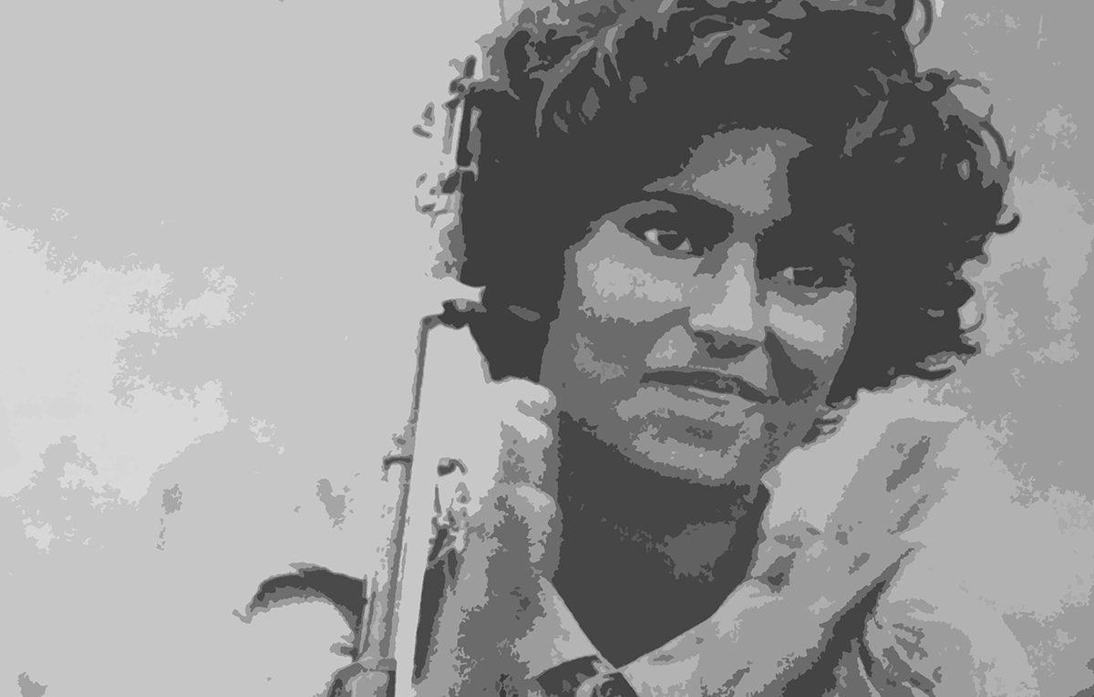 L'image montre une femme avec des cheveux bouclés, tenant un fusil. Elle sourit légèrement tout en applaudissant, ce qui semble indiquer un moment de célébration ou de camaraderie. Son uniforme militaire est visible, ce qui suggère qu'elle est impliquée dans un contexte de lutte ou de conflit. L'arrière-plan est flou, mettant l'accent sur sa figure.