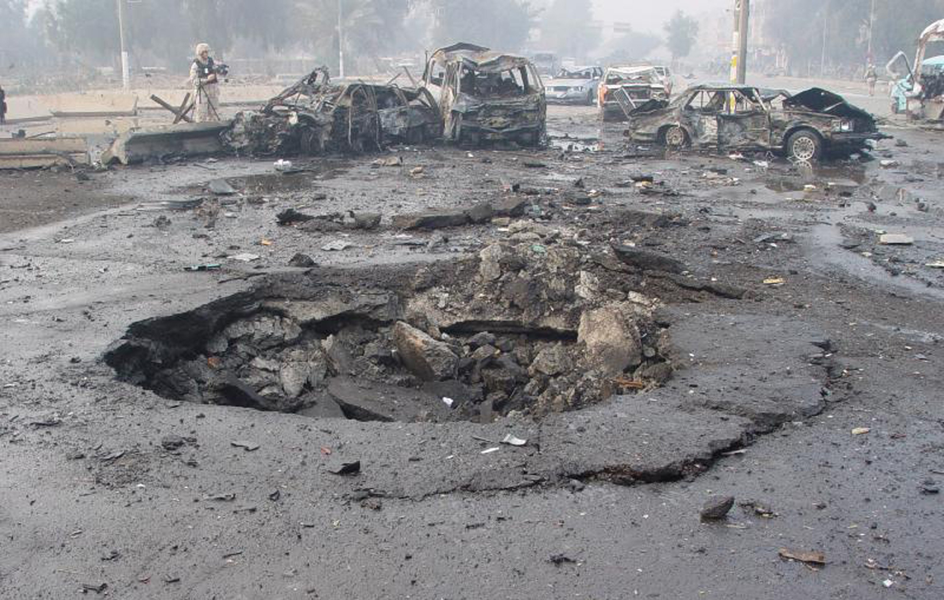 L'image montre un site de dévastation après une explosion. On peut voir plusieurs voitures endommagées et partiellement détruites, gisant sur le sol. Au centre, un cratère profond et large est présent dans le bitume, indiquant la force de l'explosion. L'environnement semble chaotique avec des débris éparpillés et une atmosphère troublée, probablement à cause de fumée ou de poussière. Cette scène évoque une situation d'urgence ou un acte de violence, reflétant des dommages importants.